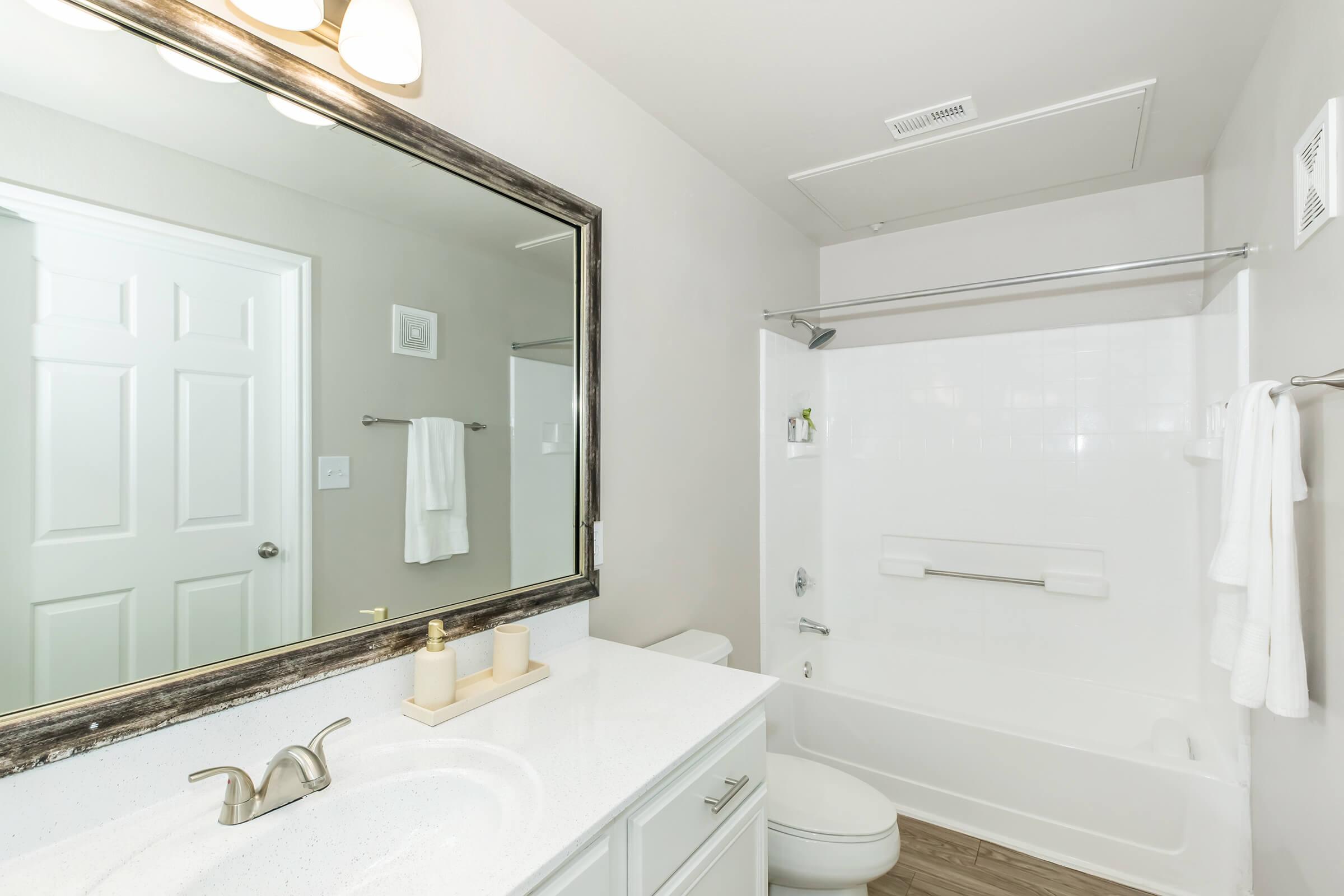 A modern bathroom with a white bathtub and shower, a large mirror above the sink, and white towels hanging on a rack. The walls are light-colored, and there is a door leading to another room. The sink area features a countertop with soap and decor items.