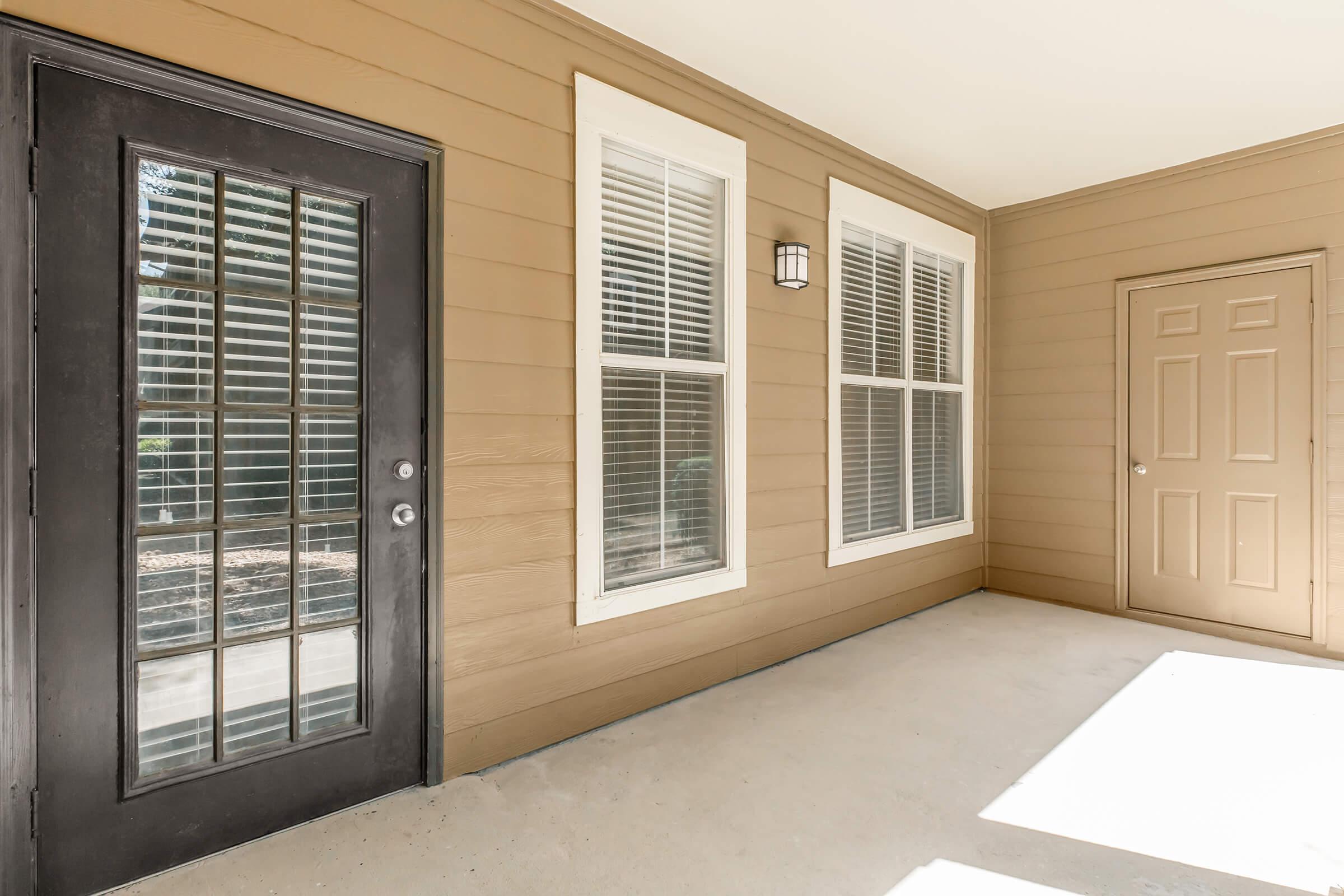 Exterior view of a well-lit porch featuring a black entry door with glass panels, two large windows with white blinds, and a beige wall. A second door is visible on the right side of the image. The porch has a clean, concrete floor and is illuminated by natural light.
