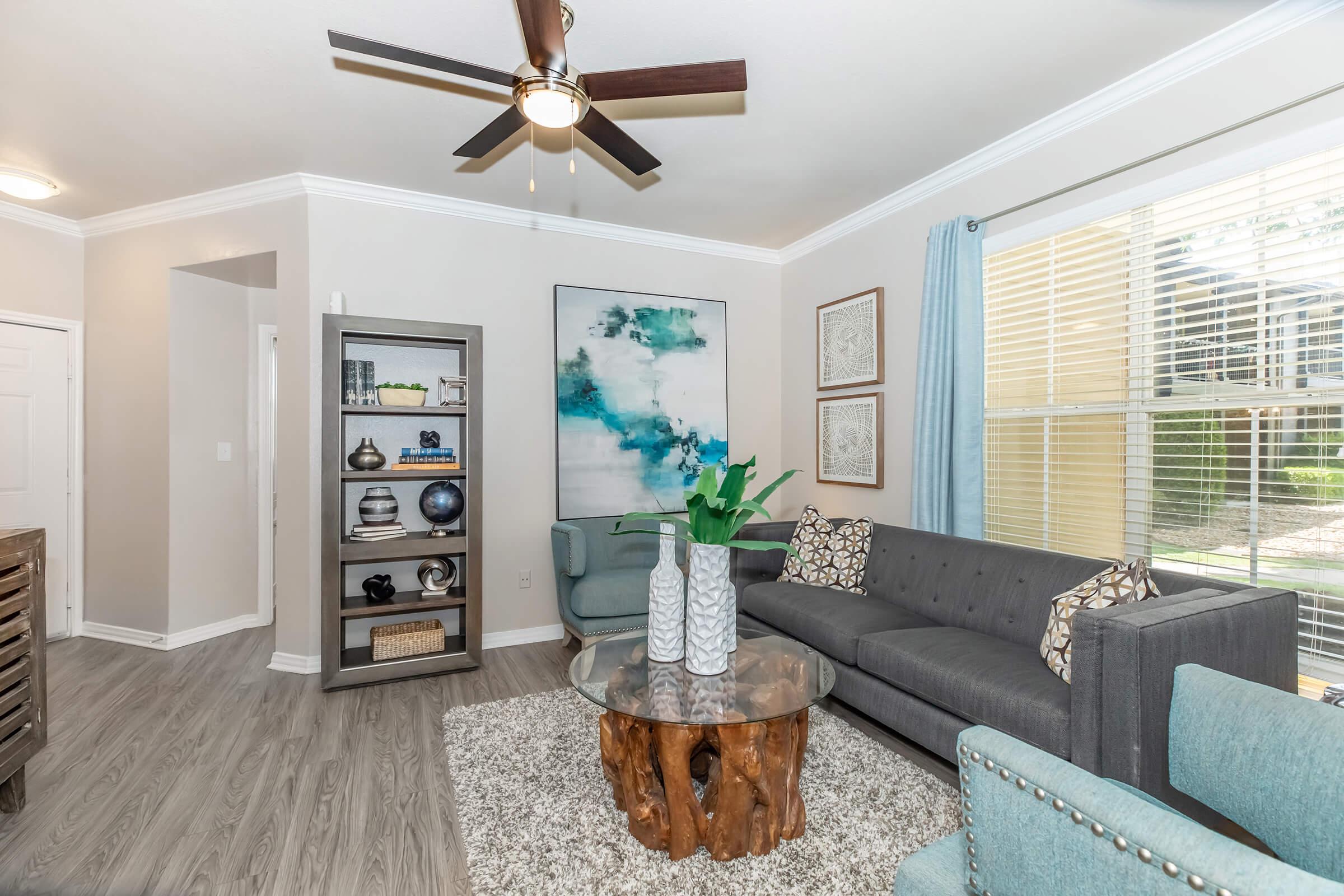 A modern living room featuring a gray sofa, a light blue accent chair, and a wooden coffee table with a textured top. A large abstract painting adorns the wall, complemented by a stylish bookshelf filled with decor. Natural light streams in through the window, creating a cozy atmosphere.