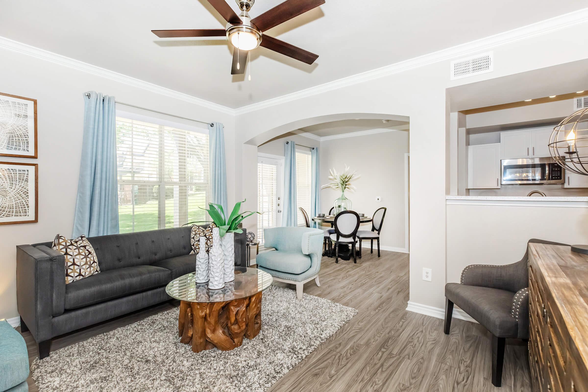 A well-lit living room featuring a gray sofa with decorative pillows, a light blue accent chair, and a wood coffee table. The space has a modern aesthetic with a ceiling fan, light-colored walls, dark wood flooring, and decorative plants. A dining area is visible in the background.