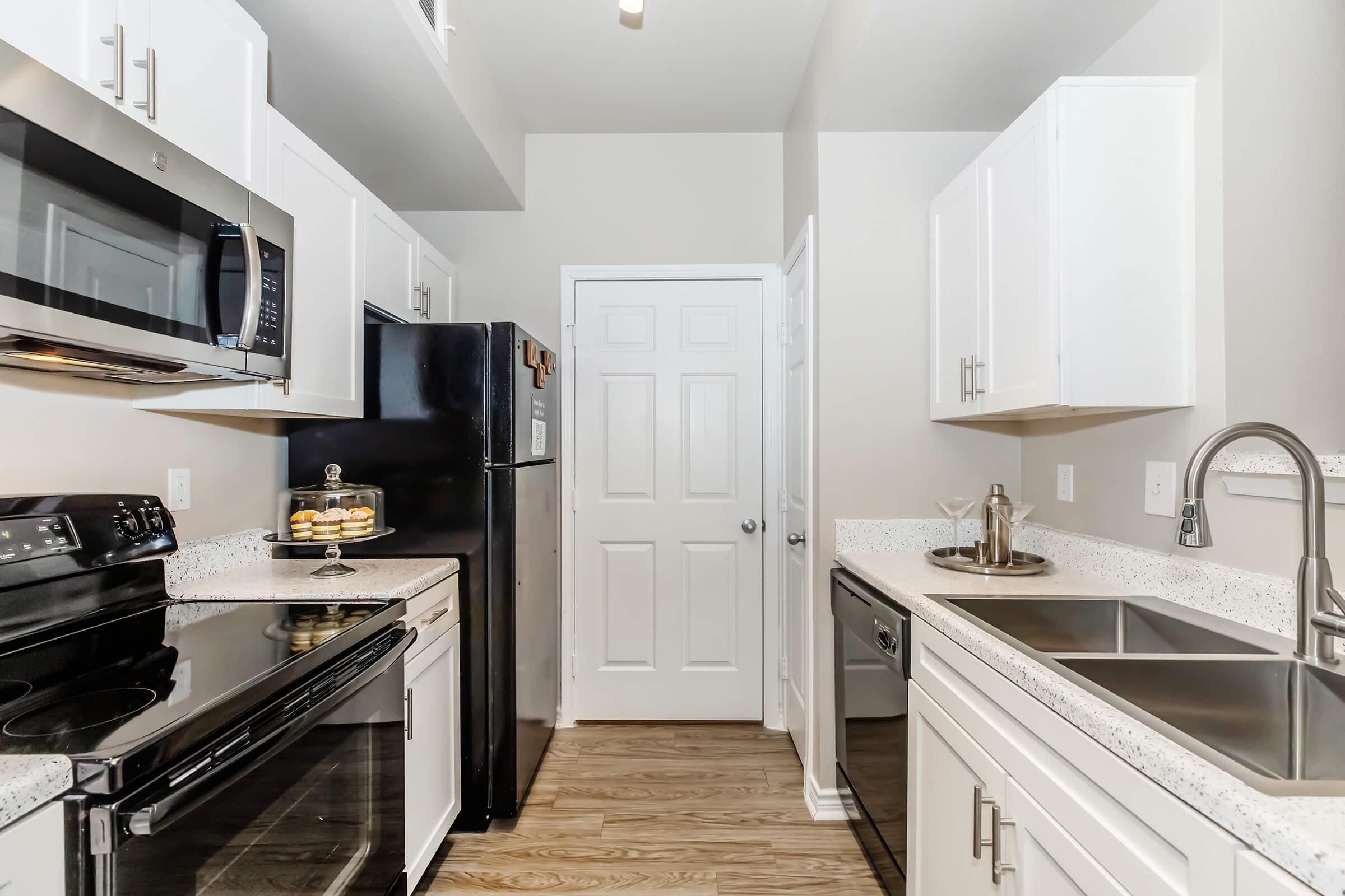 Modern kitchen featuring white cabinetry, stainless steel appliances, a black refrigerator, and a countertop with a double sink. The space is well-lit, with a door leading to another room. A glass container with cookies is prominently displayed on the countertop.