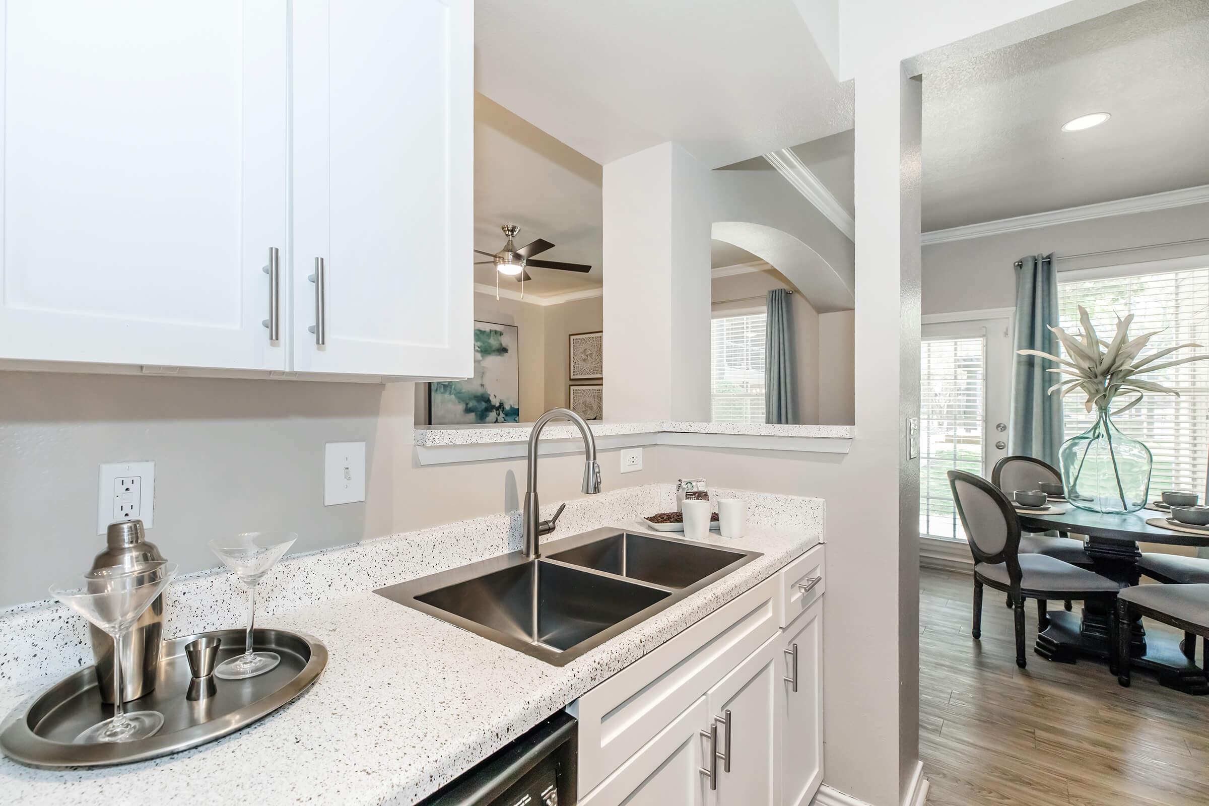 Modern kitchen with white cabinetry and quartz countertops. A stainless steel double sink features a sleek faucet. To the left, a bar cart with cocktail glasses is visible. The background shows a dining area with a table set for two, and large windows allowing natural light to brighten the space.