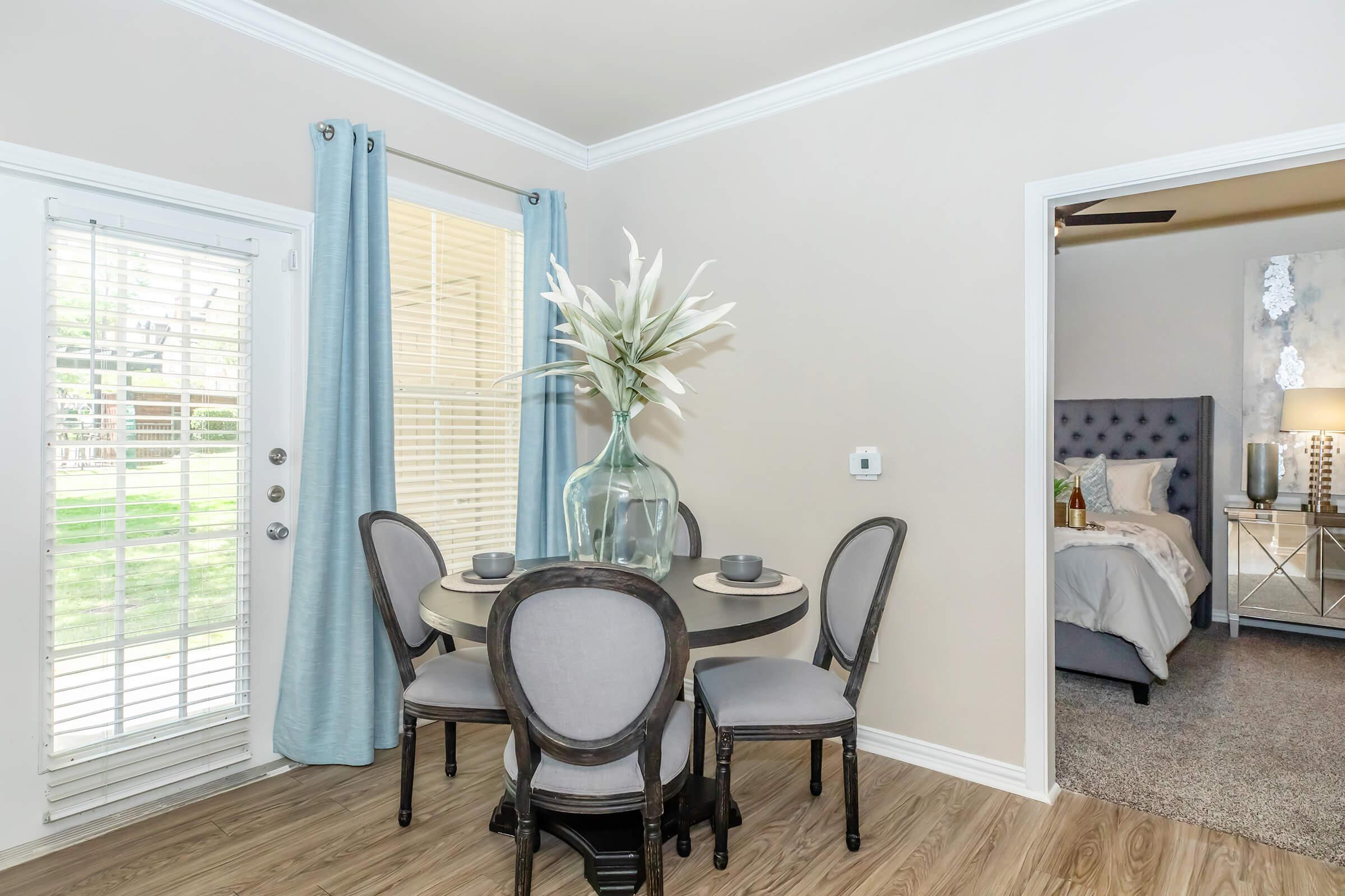 A cozy dining area featuring a round table with two plates and cutlery. Four elegant chairs surround the table, with a large vase of white lilies as the centerpiece. Light blue curtains frame a window, allowing natural light to fill the space. In the background, a bedroom is visible through an open door.