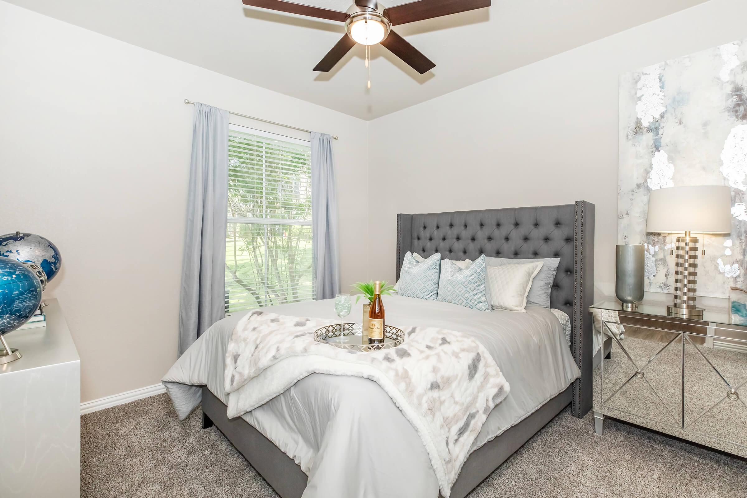Cozy bedroom featuring a tufted gray bed with soft bedding, decorative pillows, and a tray on the bed. A tall lamp with a reflective base stands beside a stylish nightstand. The room has light-colored walls, a window with sheer curtains, and plush carpet, creating a warm and inviting atmosphere.