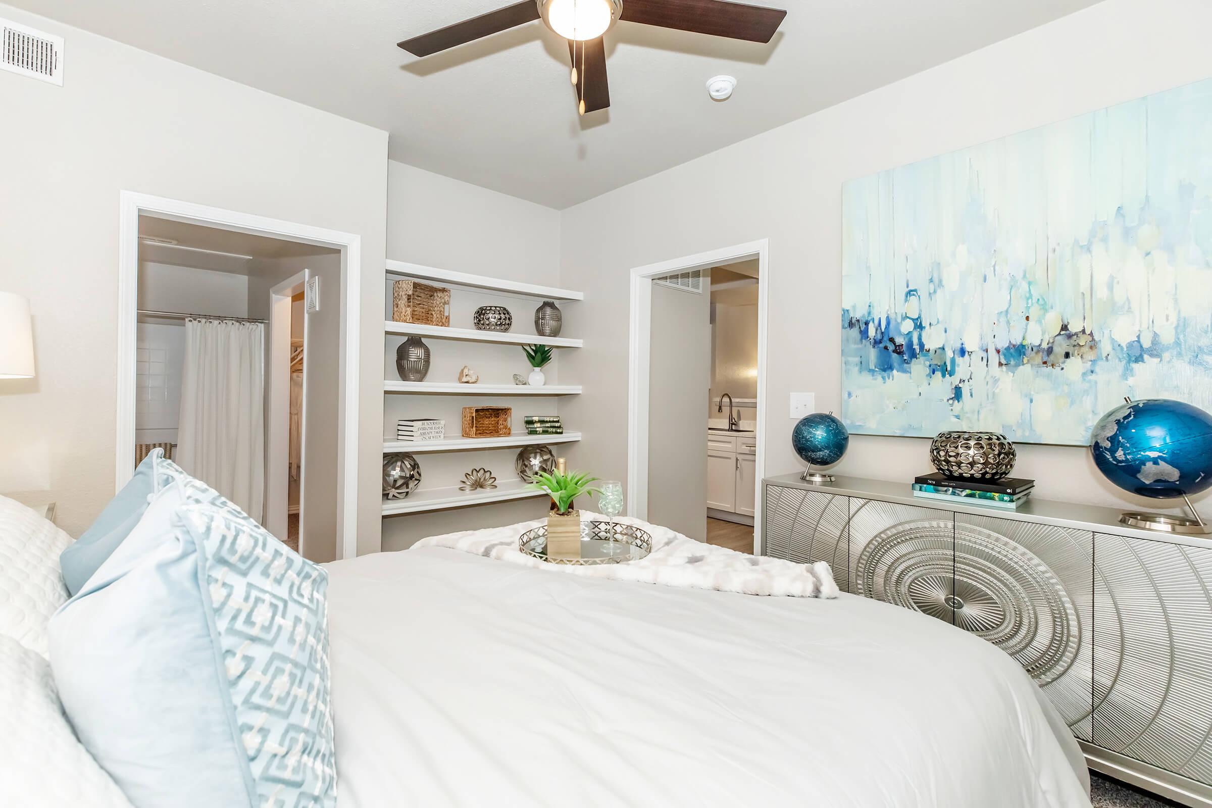 A cozy bedroom featuring a large bed with white linens and decorative pillows. There are shelves with decorative items, and a stylish chest of drawers with a blue globe and a plant. The walls are painted in soft tones, and a piece of abstract art hangs above the chest. A doorway leads to a bathroom, and another opens to a hallway.