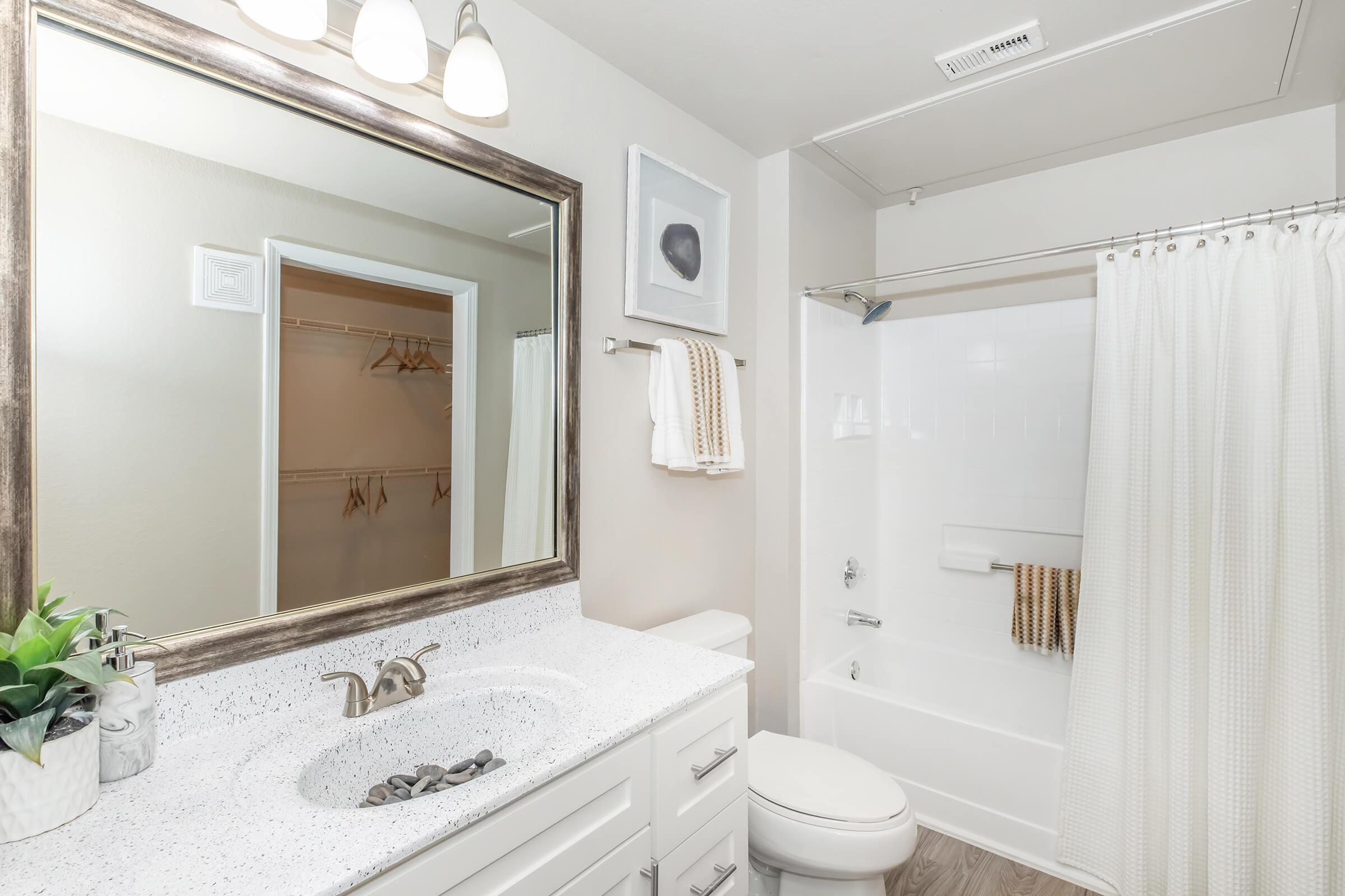 A modern bathroom featuring a large mirror with a decorative frame, a white countertop with a small plant, and a shower area with a white curtain. The walls are painted in a light color, and there's a towel hanging on a rack. A cabinet with an open closet is visible in the background.
