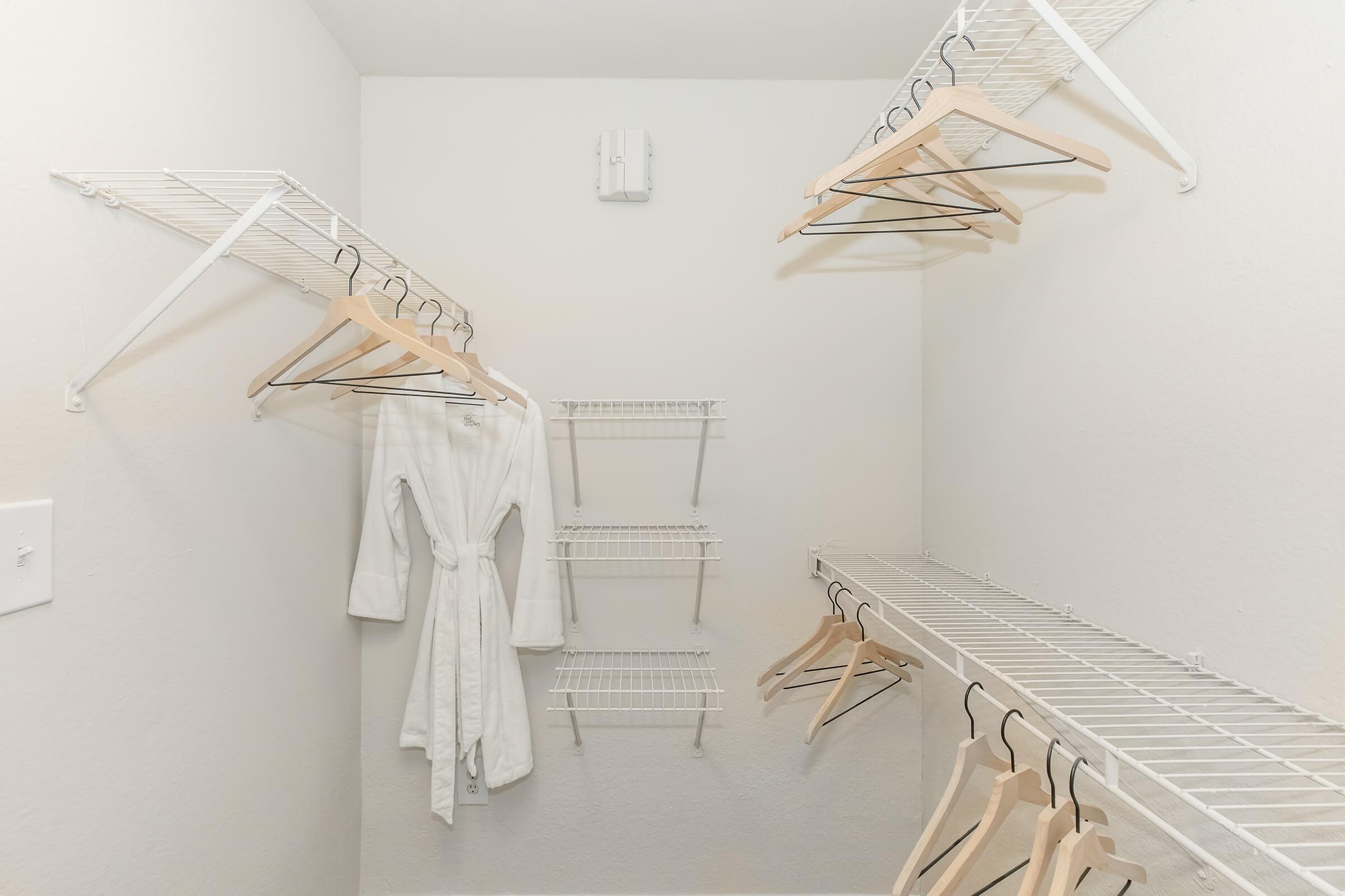 Empty closet with white walls and wire shelving. A white bathrobe hangs on one of the hangers, while several empty hangers are arranged on the shelves. The space is organized and well-lit, ideal for clothing storage.