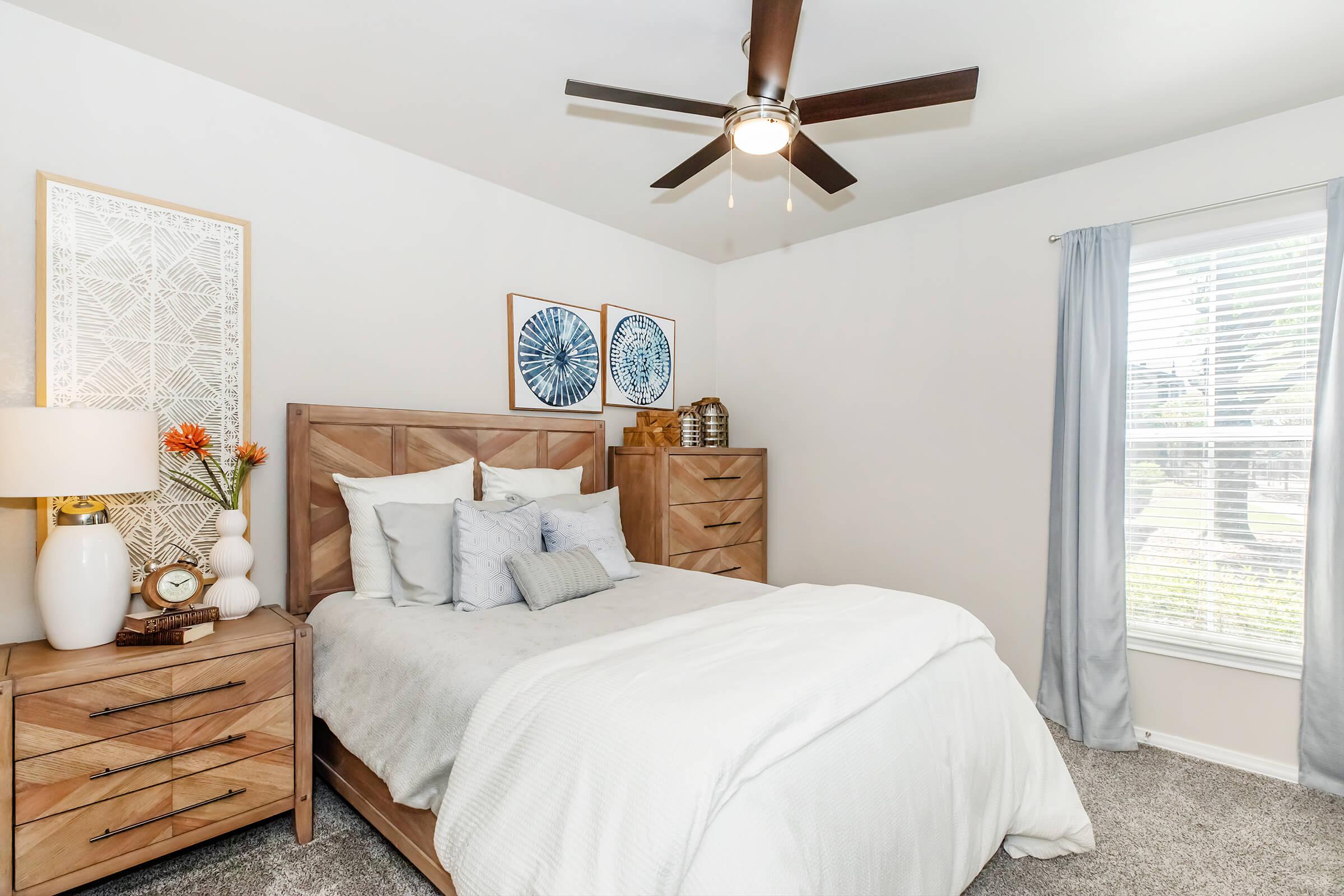 A cozy bedroom featuring a wooden bed with white bedding, a nightstand with a lamp and decorative items, and two wall art pieces. A ceiling fan is above the bed, and there's a window with sheer curtains allowing natural light to enter the room.