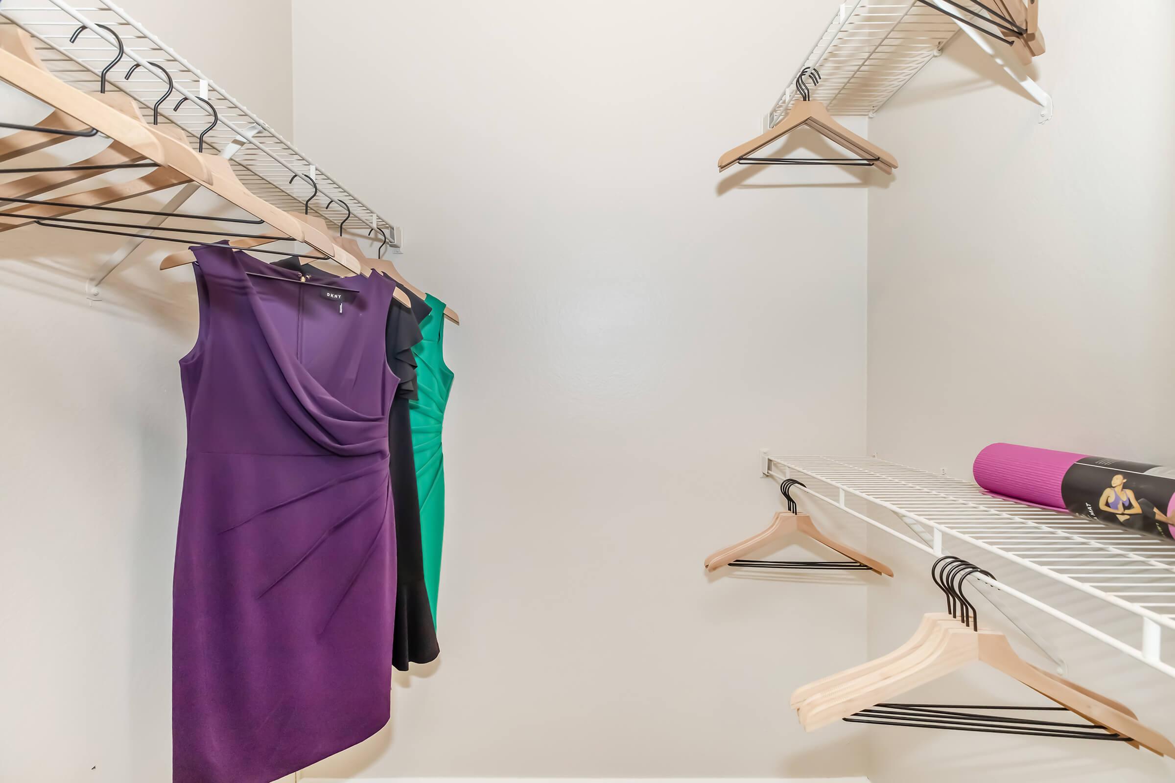 Two dresses hanging in a well-organized closet: one purple dress on the left and one green dress on the right. There are empty hangers on the left side and a shelf on the right, with a rolled-up yoga mat placed on the top shelf. The closet walls are painted white.