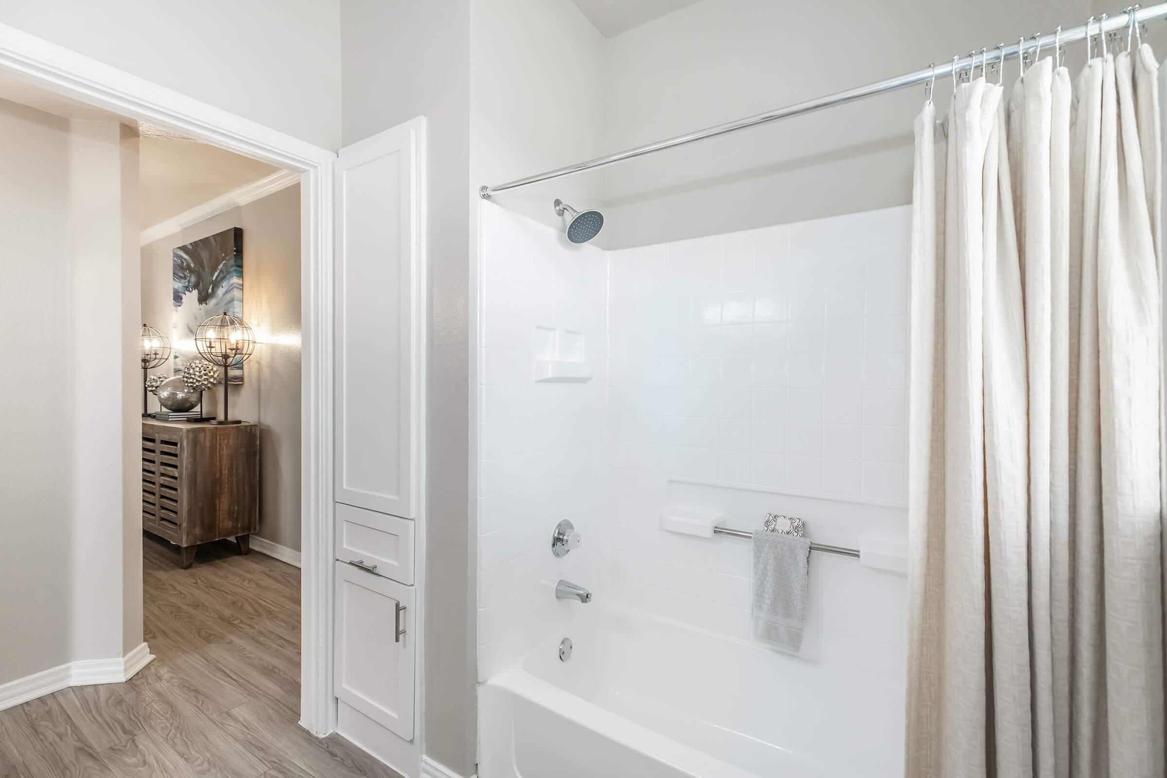 A clean and modern bathroom featuring a white bathtub with a shower curtain, a shower head mounted above, and a light-colored floor. In the background, a doorway leads to another room with stylish decor and soft lighting.