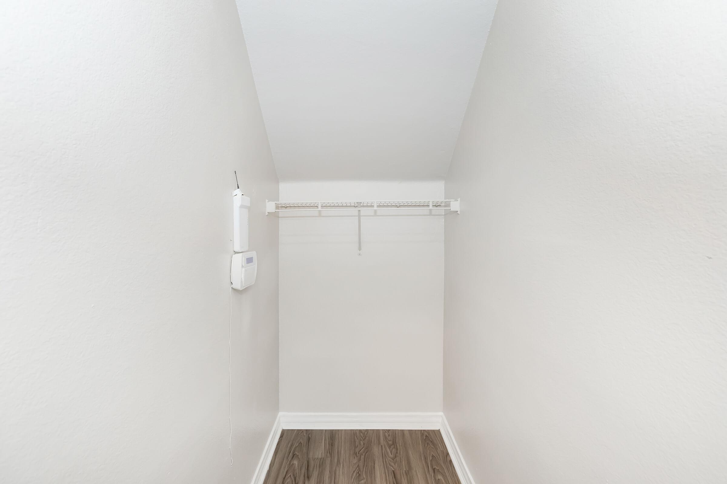 An empty, well-lit closet with white walls and a sloped ceiling. There is a single shelf along the back wall, and the floor features a wood-like finish. A small device is mounted on the wall near the entrance.