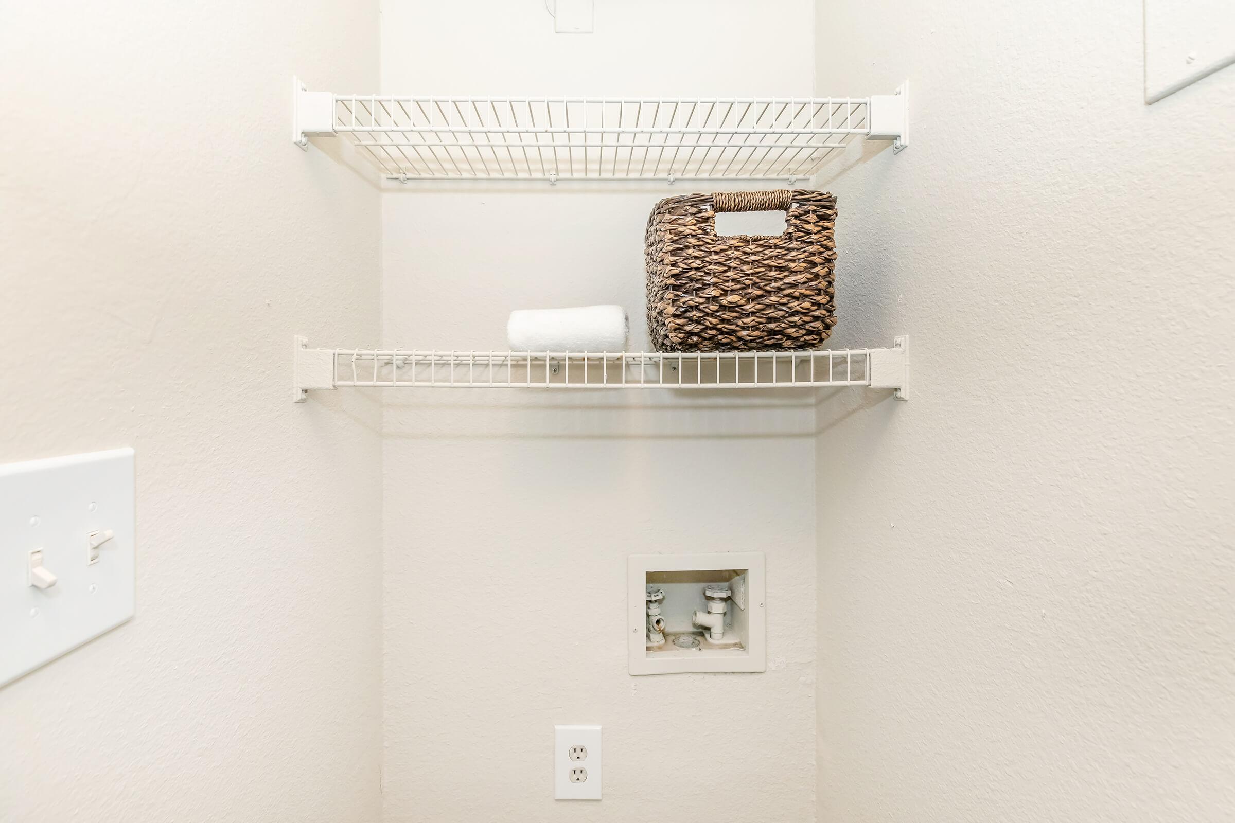 A small storage space featuring two wire shelves. The top shelf holds a woven basket, while the bottom shelf has a rolled towel. A small access panel for plumbing is visible on the wall, along with light switches and an electrical outlet. The walls are painted a light neutral color.