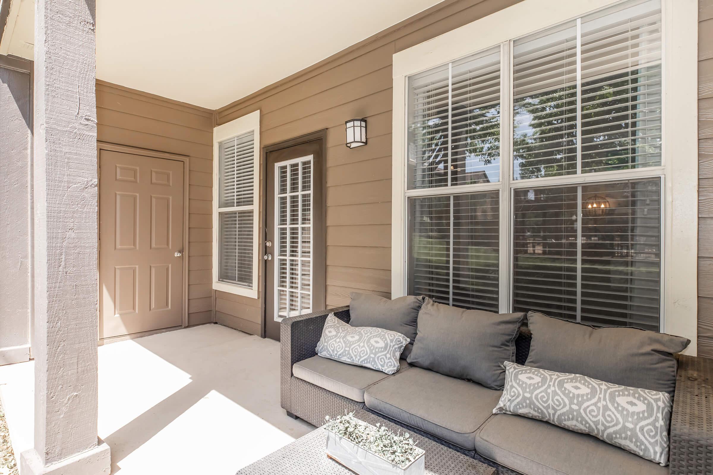 A cozy outdoor patio area featuring a comfortable gray couch with decorative pillows, a small table with a plant, and a door with a light fixture above. The walls are tan-colored with large windows and shutters, enhancing the inviting atmosphere. Natural light streams in, creating a warm ambiance.