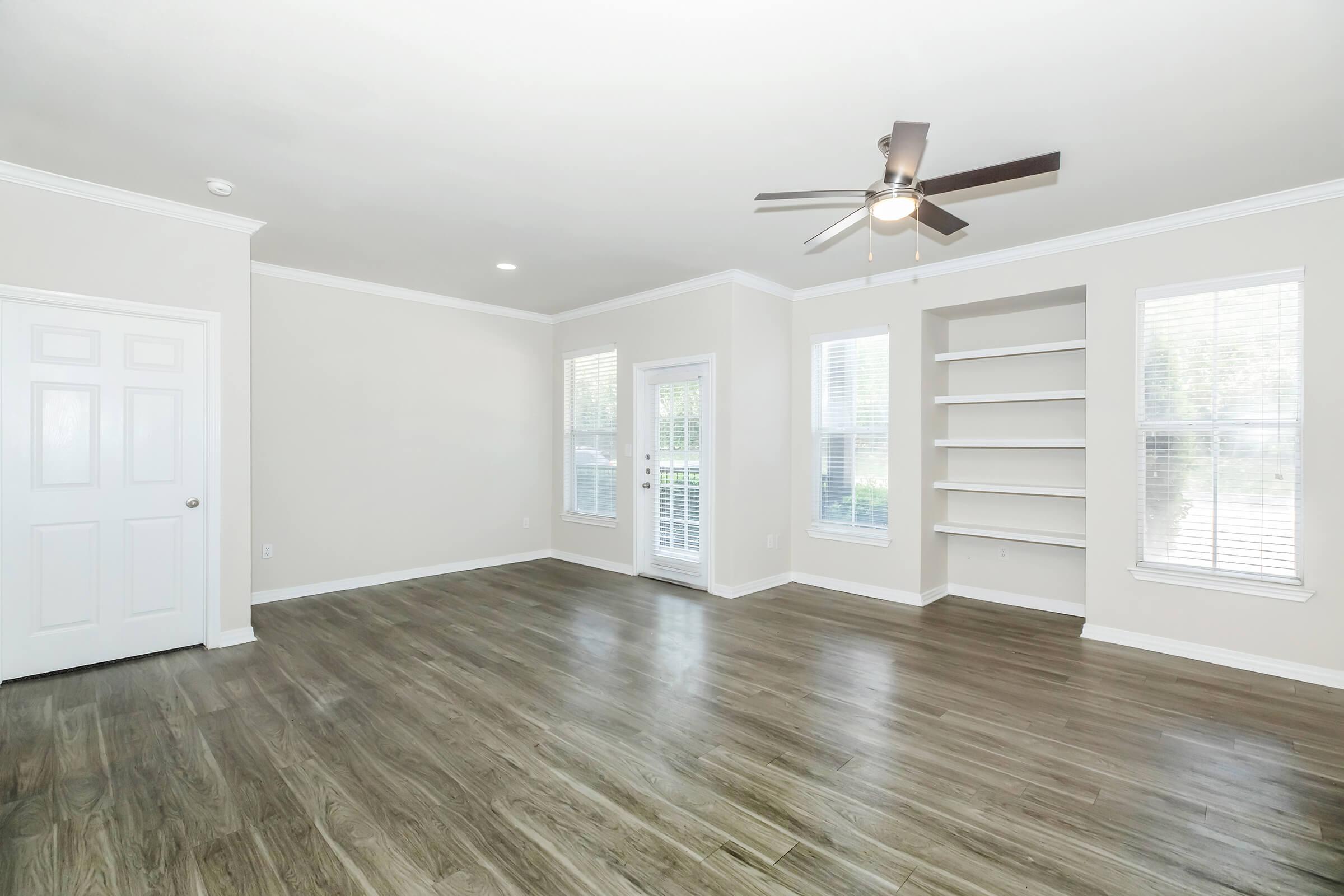 Spacious, empty living room with light-colored walls and laminate flooring. Features a ceiling fan, large windows allowing natural light, and a door leading outside. Includes built-in shelves on one side and a closet door on the left. Ideal for personal decoration and furniture arrangement.