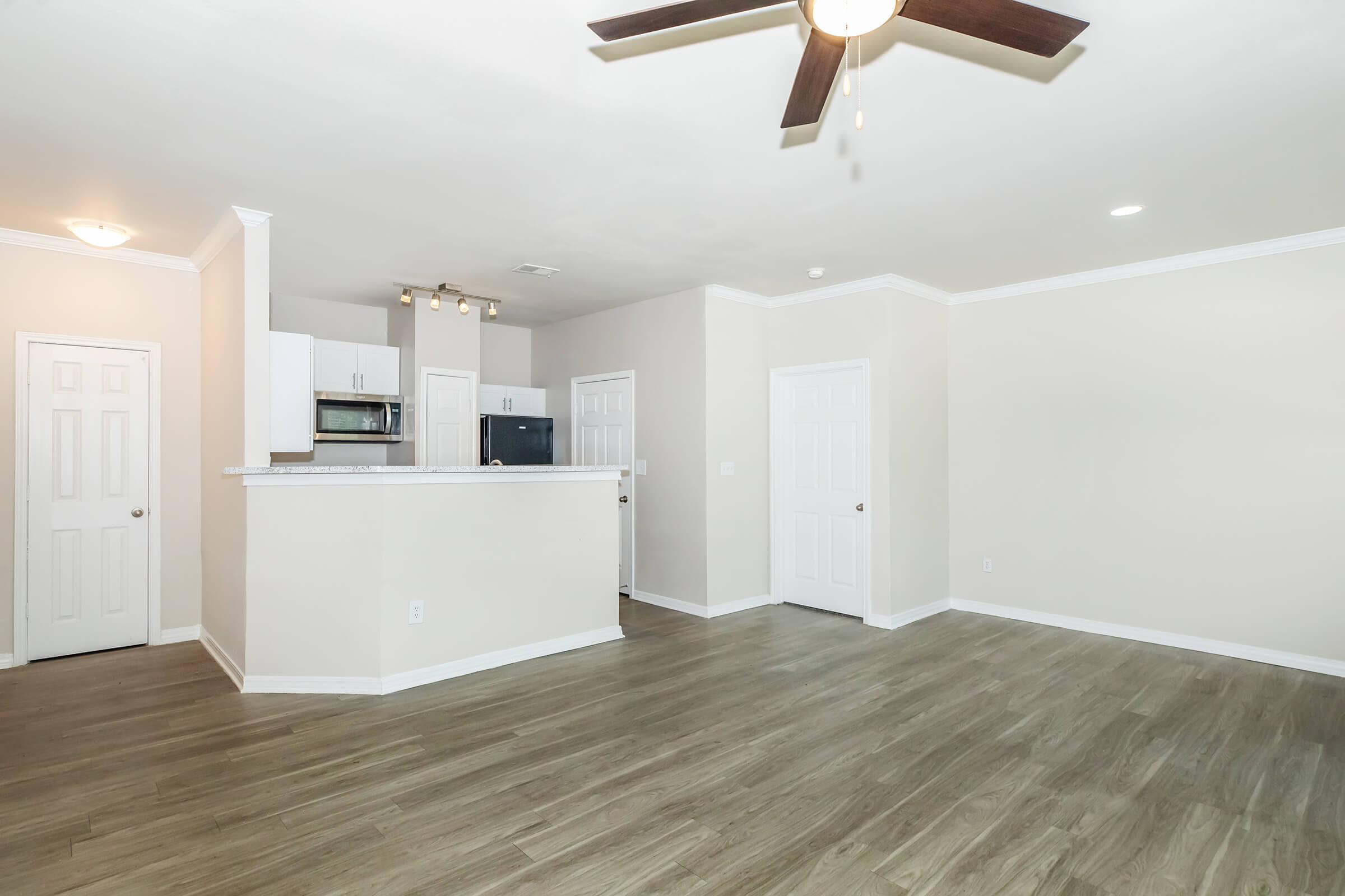 A spacious living area with light-colored walls and a ceiling fan. The room features a kitchen area with a breakfast bar, a refrigerator, and an open layout. Two doors lead to other rooms, and the flooring is a light wood finish. Natural light brightens the space.