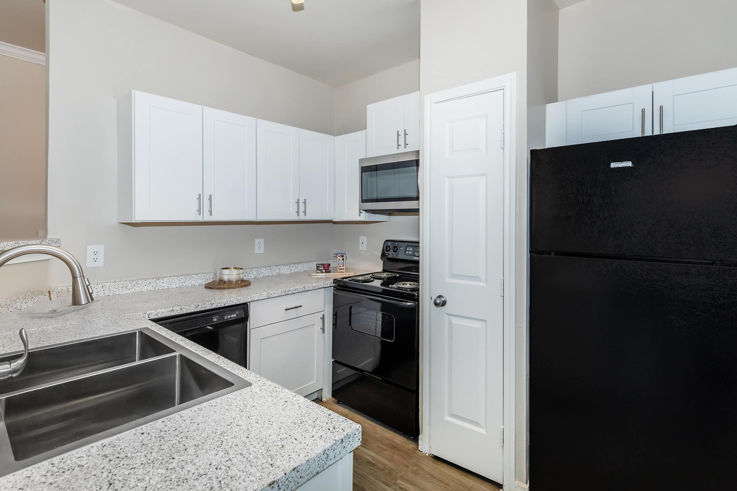 A modern kitchen featuring white cabinets, stainless steel appliances, a black refrigerator, and a double kitchen sink. The countertops are light-colored with a speckled pattern, and the space is well-lit with natural light. A door leads to another room, enhancing the kitchen's accessibility and functionality.
