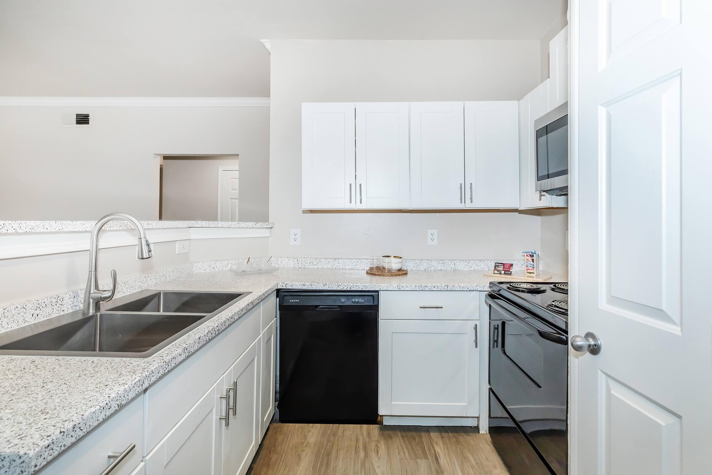 Modern kitchen featuring white cabinets, granite countertops, a double sink, and stainless-steel appliances including a dishwasher and stove. The interior is bright and airy, with light-colored walls and wooden flooring, creating a clean, contemporary aesthetic.
