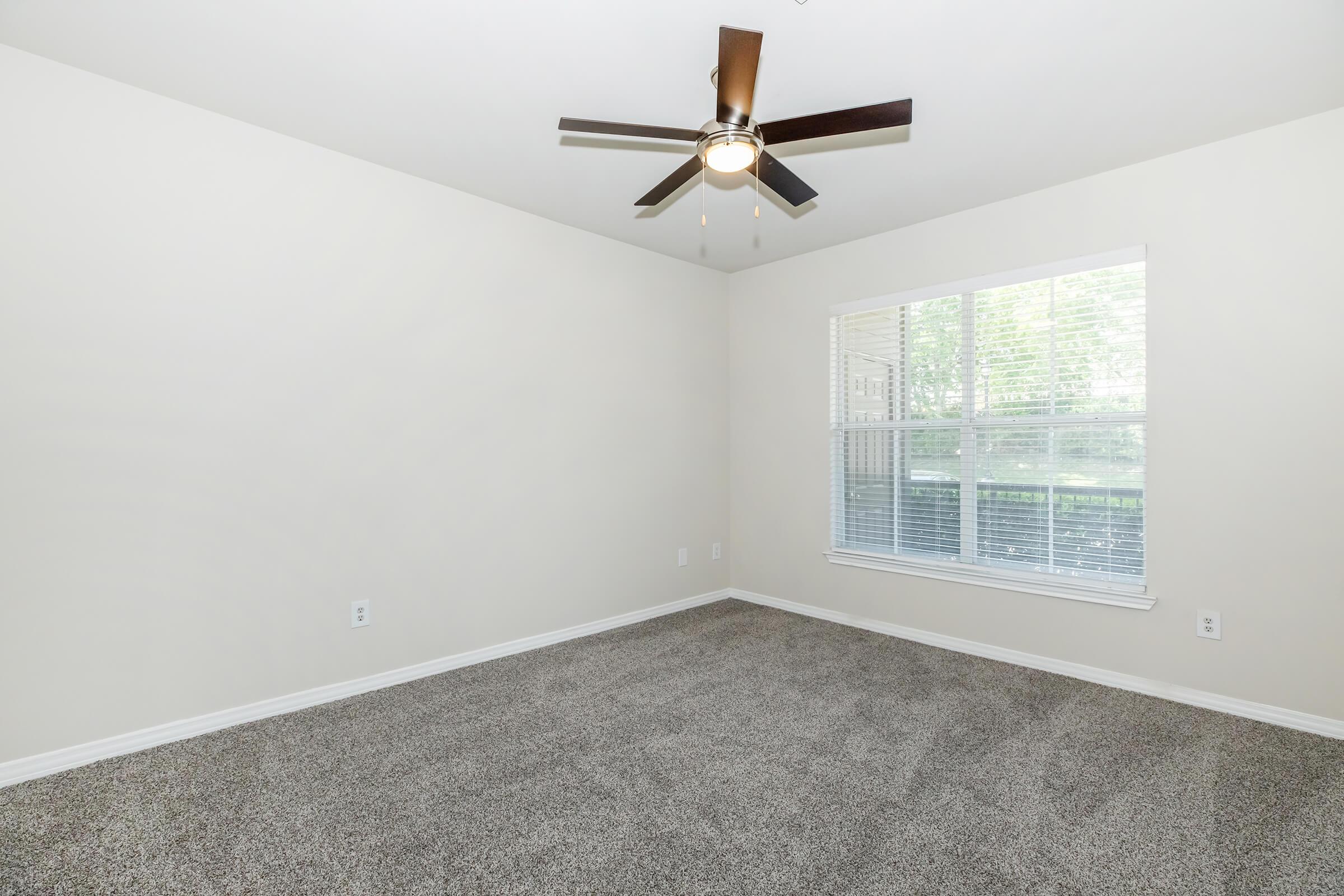 A well-lit, empty room with light beige walls and a large window featuring blinds. The floor is covered in gray carpet, and a ceiling fan with dark blades is installed in the center. The space appears clean and modern, ready for furniture.