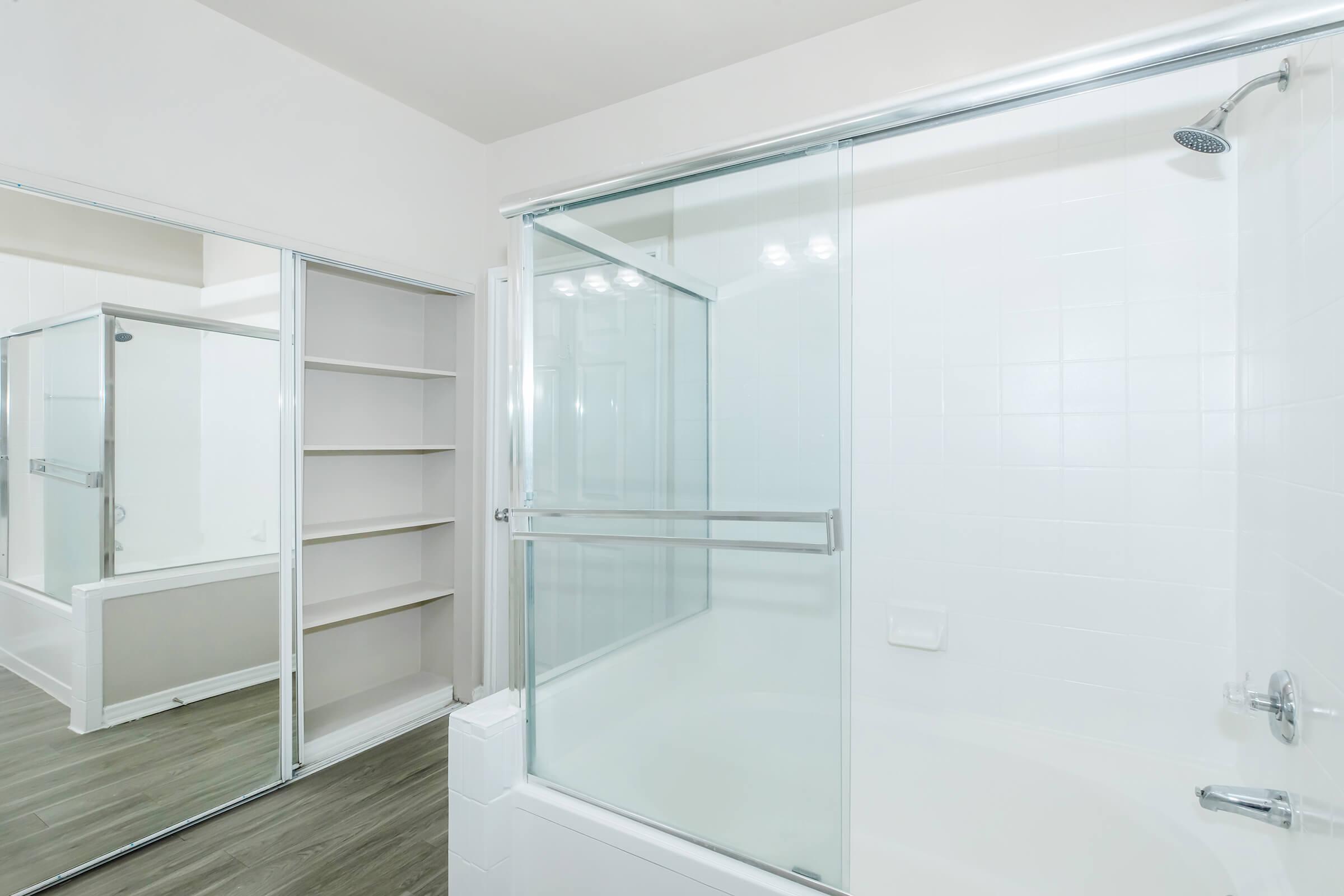 A modern bathroom showcasing a glass shower enclosure with a sleek design, gray wood-look flooring, and a built-in shelving unit adjacent to the shower area. The walls are painted in a light neutral color, creating a spacious and airy feel.