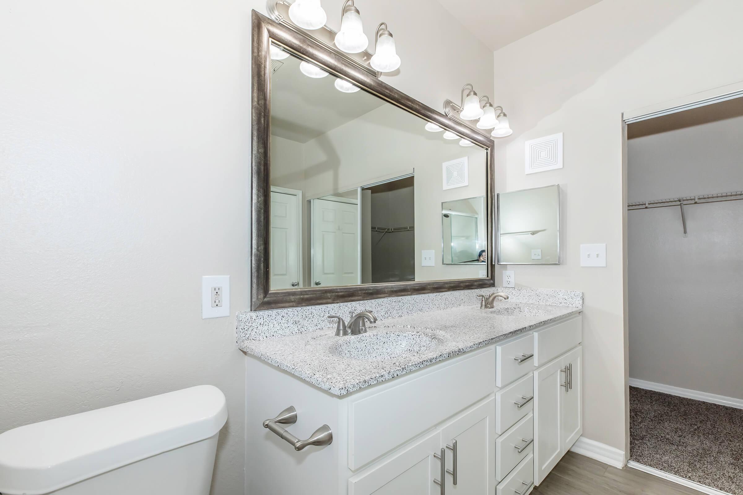 A modern bathroom featuring a double vanity with a granite countertop and two sinks, above which is a large rectangular mirror. The walls are painted a light color, and there is a well-lit area with multiple light fixtures. A closet door is visible in the background, and the flooring is a neutral shade.