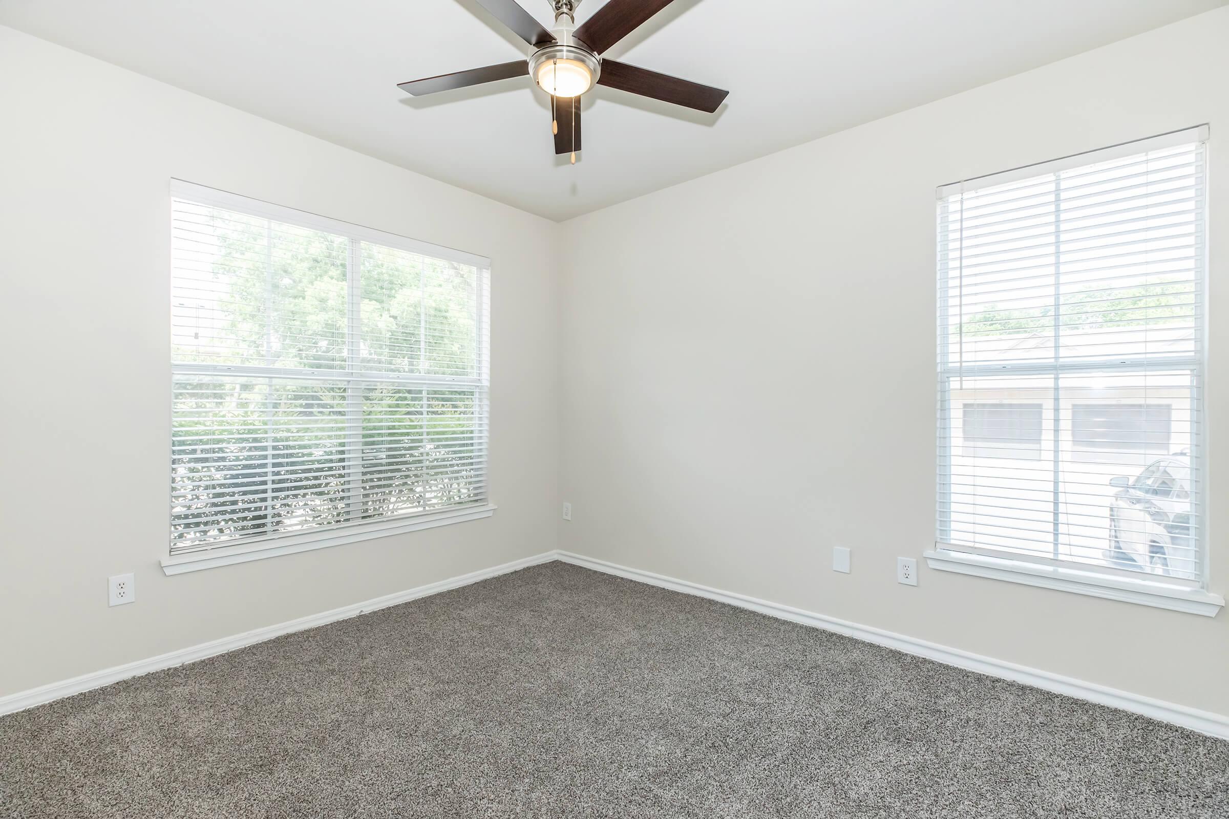 Empty room with light-colored walls and carpet. Two windows with white blinds allow natural light to enter, and a modern ceiling fan hangs from the ceiling. The room is uncluttered, providing a spacious feel, ideal for various uses such as an office or bedroom.