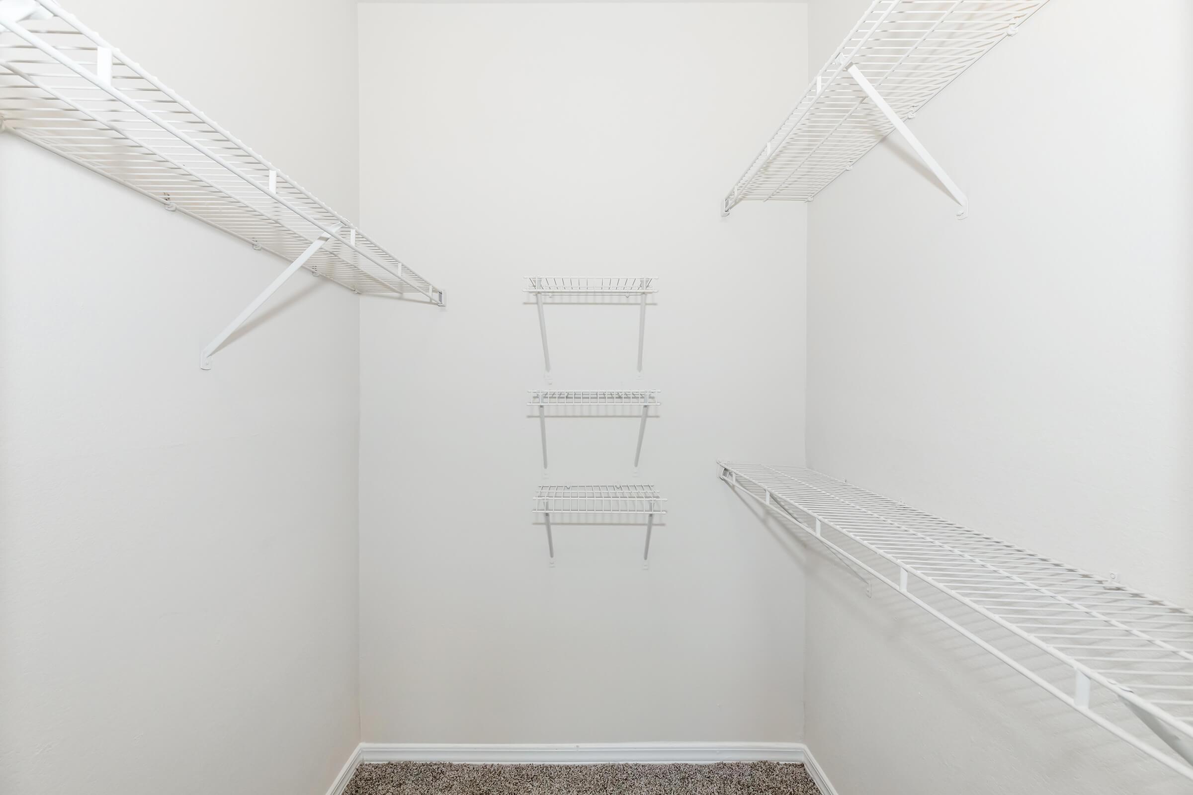 A clean, empty closet with white wire shelves mounted on light-colored walls. The floor is carpeted, and there are no items stored on the shelves. The overall appearance is minimalistic and organized.