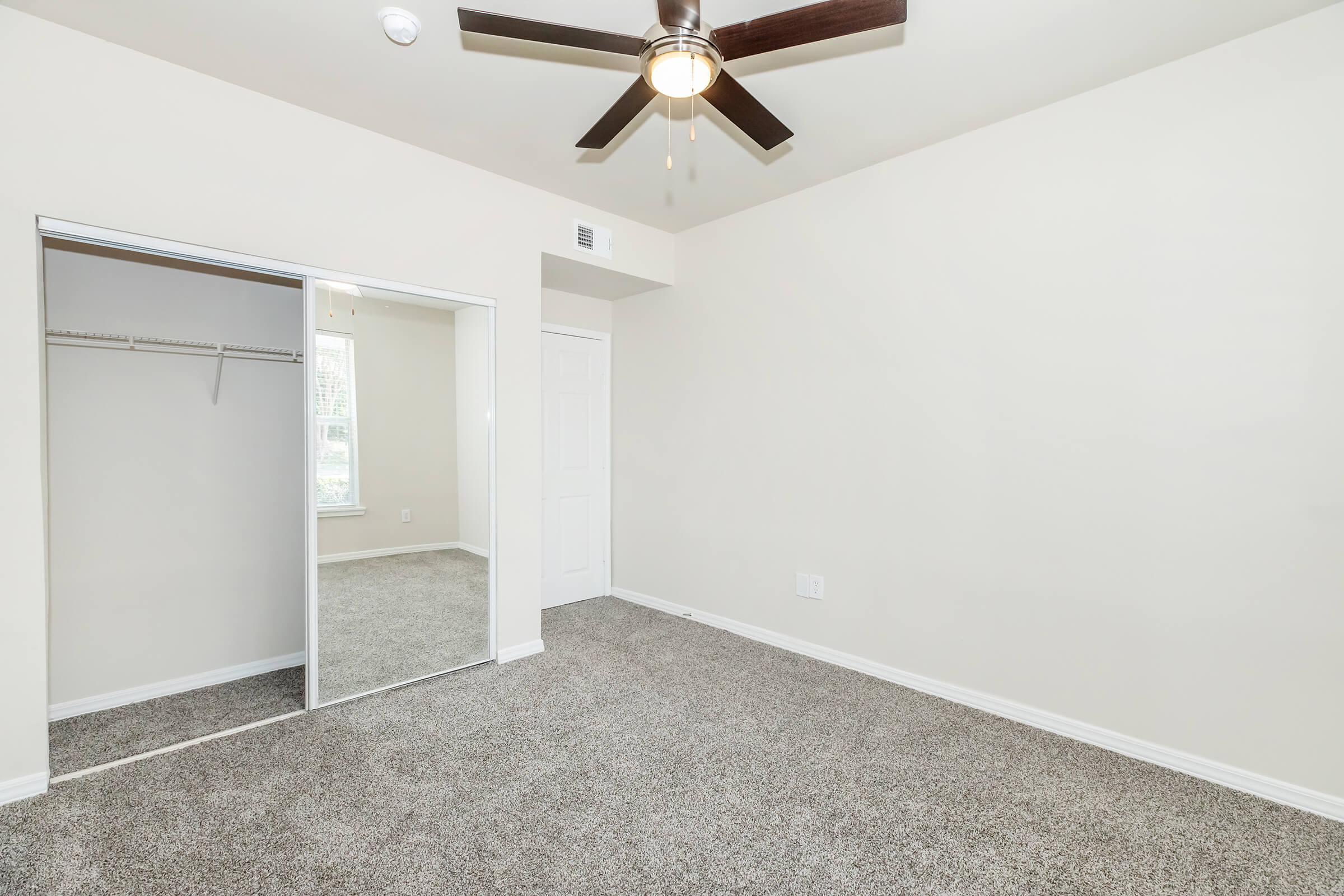 A spacious, empty room featuring light beige walls, a ceiling fan, and plush carpet. A mirrored closet door reflects the interior, and a window provides natural light. The overall ambiance is bright and inviting, suitable for various personal or decorative styles.