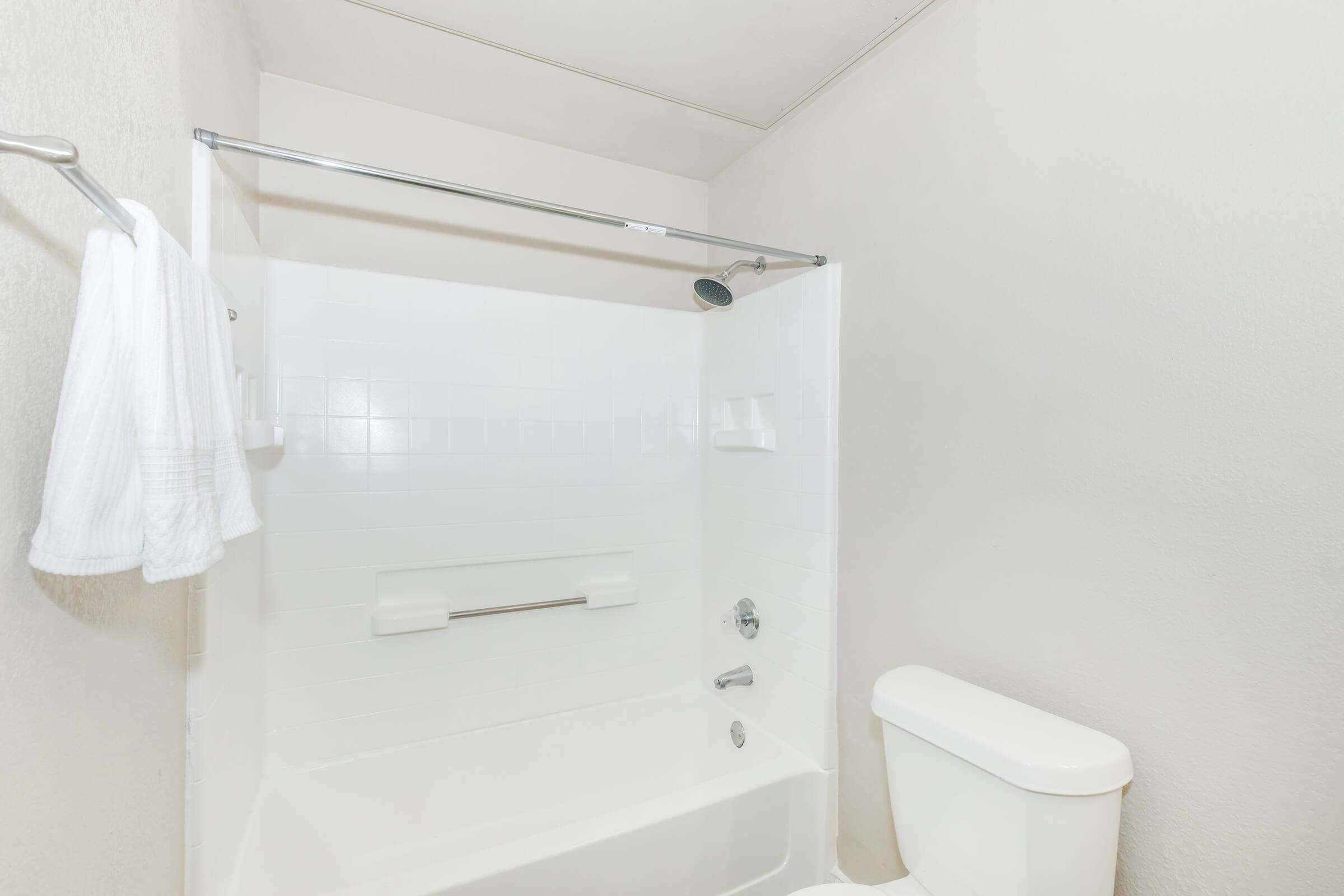 A clean, simple bathroom featuring a white tiled shower area with a silver showerhead and a bathtub. A towel hangs on a rack nearby, and there is a white toilet to the right. The walls are plain and light-colored, creating a minimalist look.