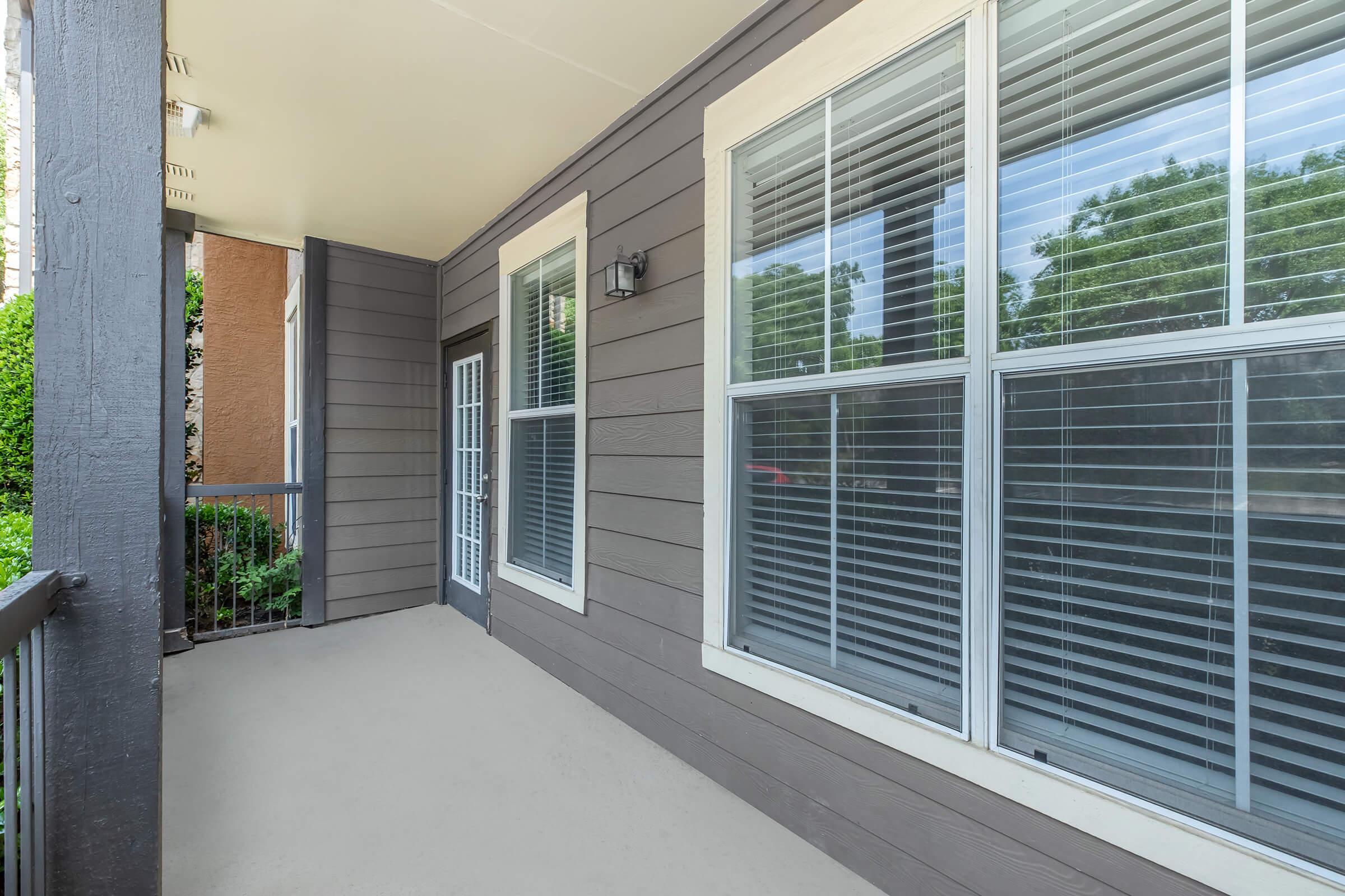 A covered porch with light gray walls and large windows featuring white blinds. The flooring is light-colored, and there is a railing along one side. Green foliage is visible outside the windows, creating a serene outdoor atmosphere.