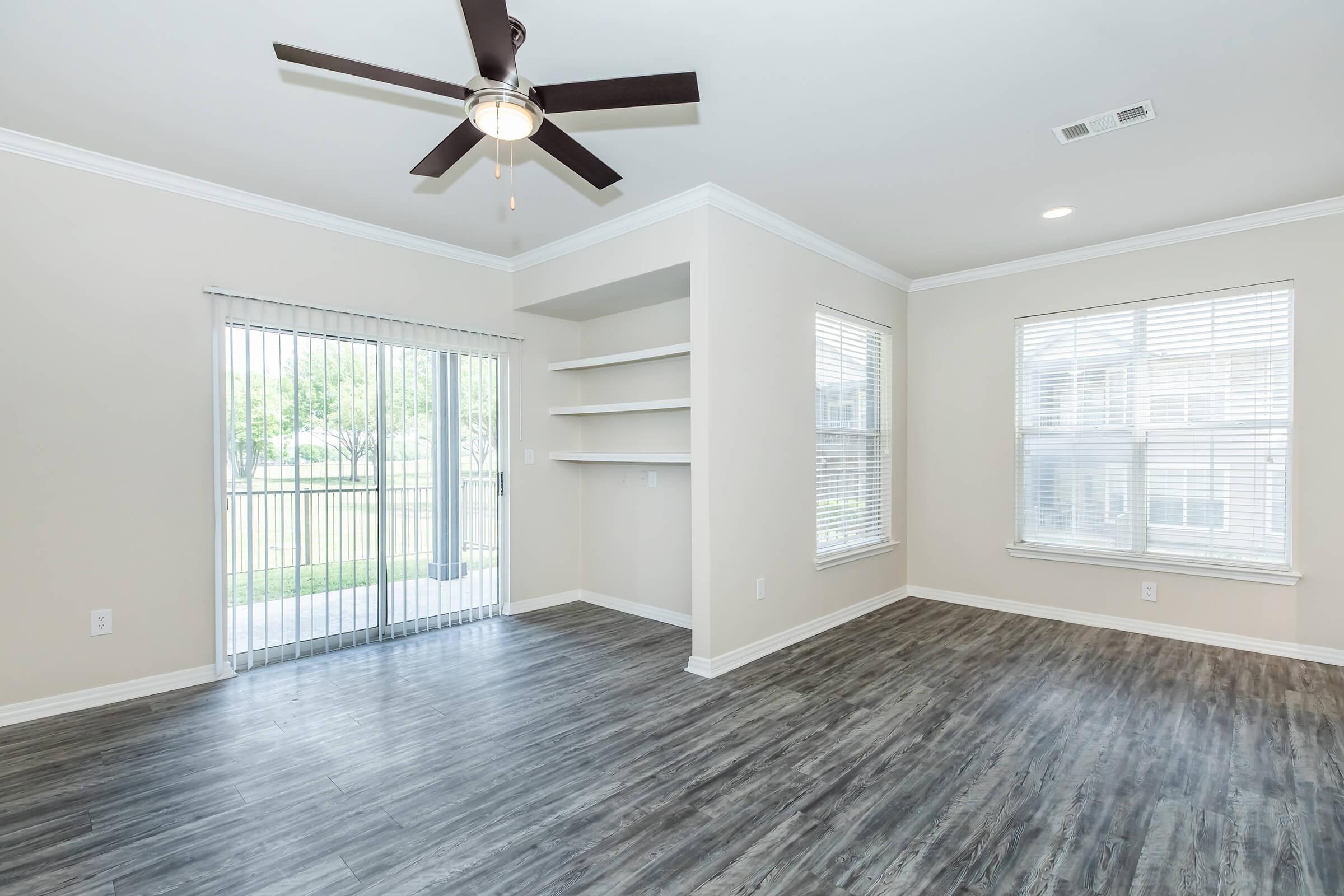 A bright, airy living room featuring a ceiling fan, light-colored walls, and wood-style flooring. It includes a sliding glass door leading to an outdoor space, built-in shelving, and multiple windows allowing natural light to fill the room.