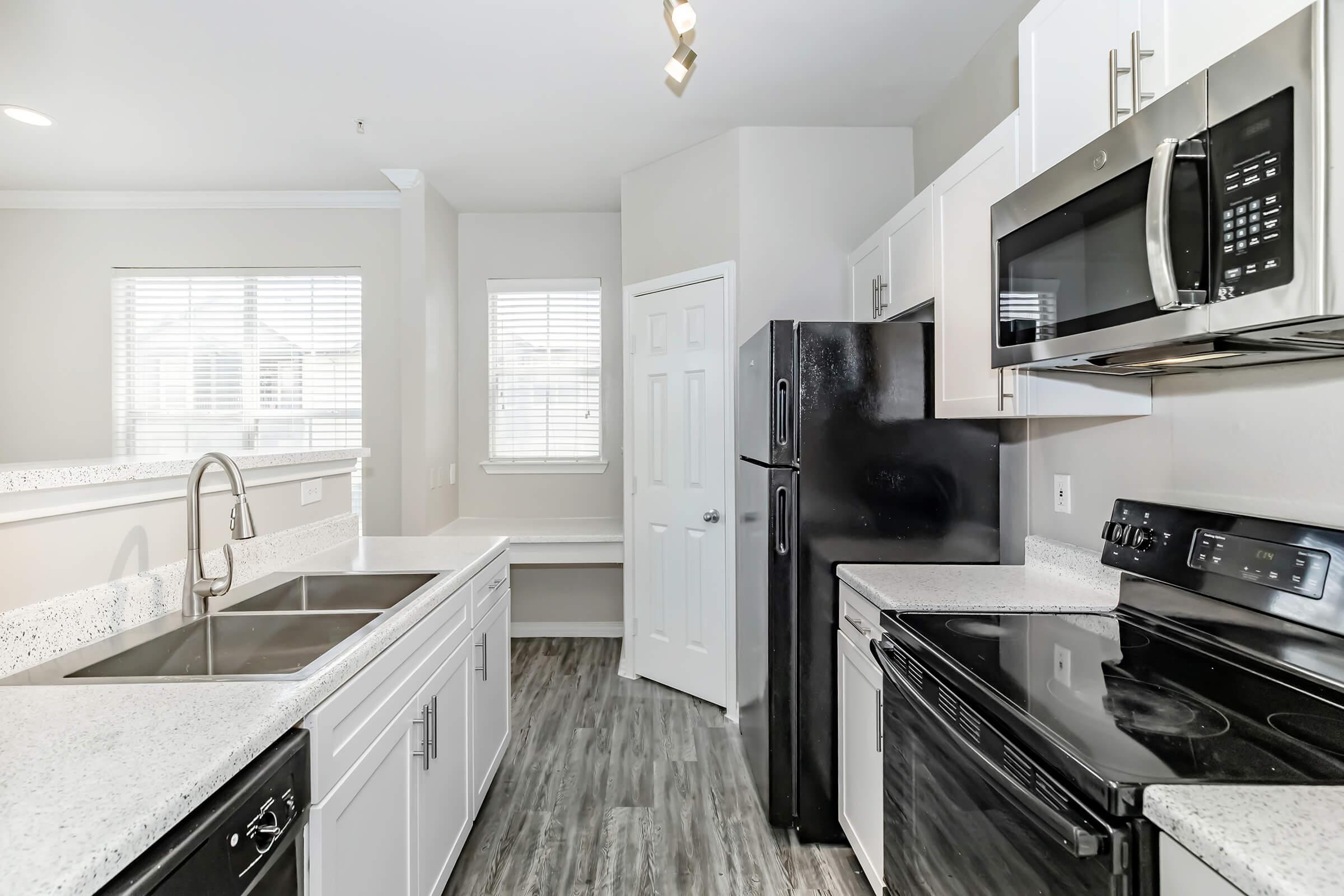 A modern kitchen featuring white cabinets, a black refrigerator, and stainless steel appliances. The countertop is speckled white with gray accents, and there are large windows providing natural light. A pantry door is visible, and the flooring is a light wood-like material.