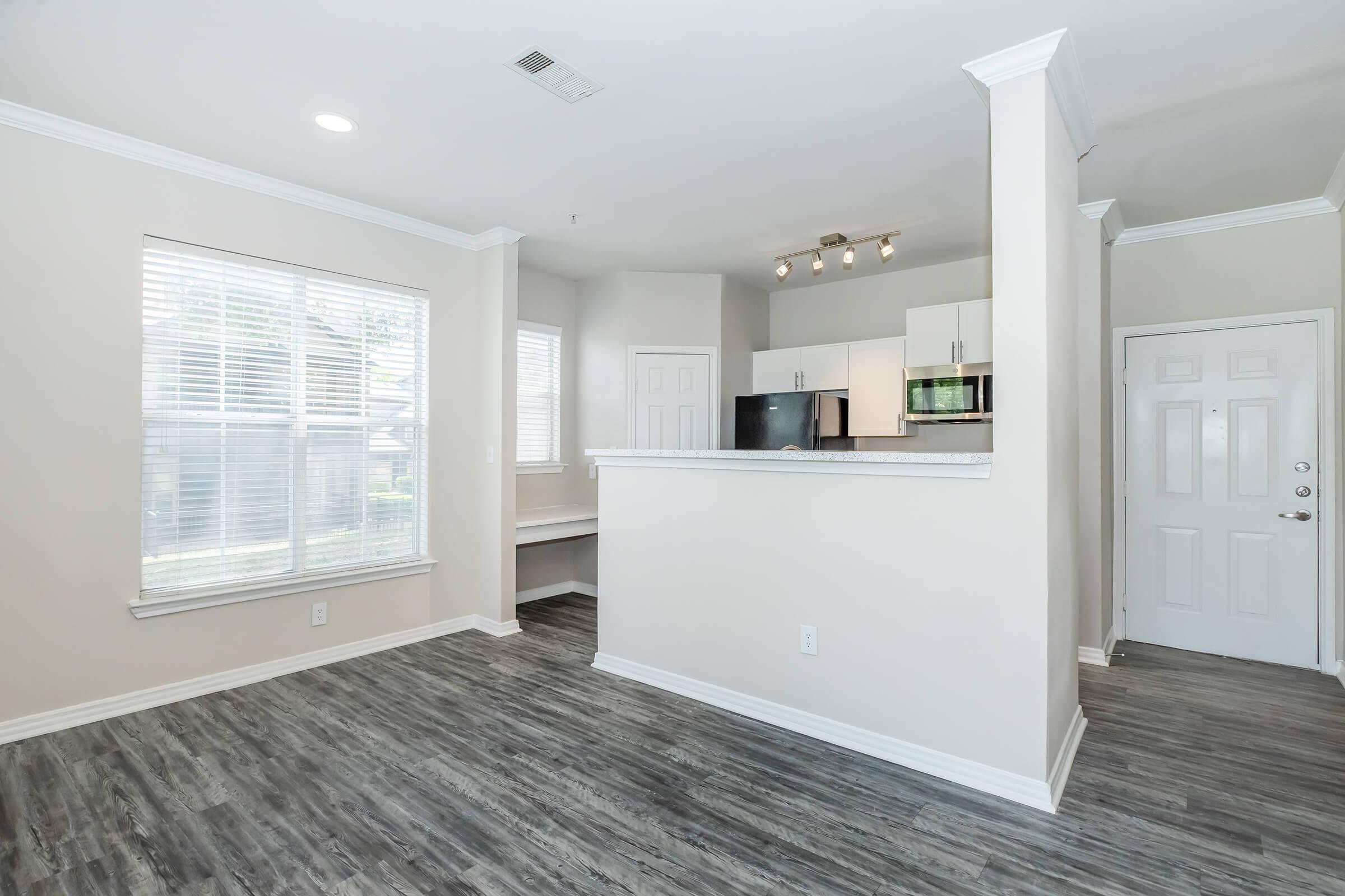 Bright, modern interior of an apartment featuring an open layout. The space includes a living area with a large window, a kitchen with white cabinetry and a granite countertop, and a doorway leading to the entrance. The flooring is a sleek, dark laminate, adding to the contemporary feel of the room.