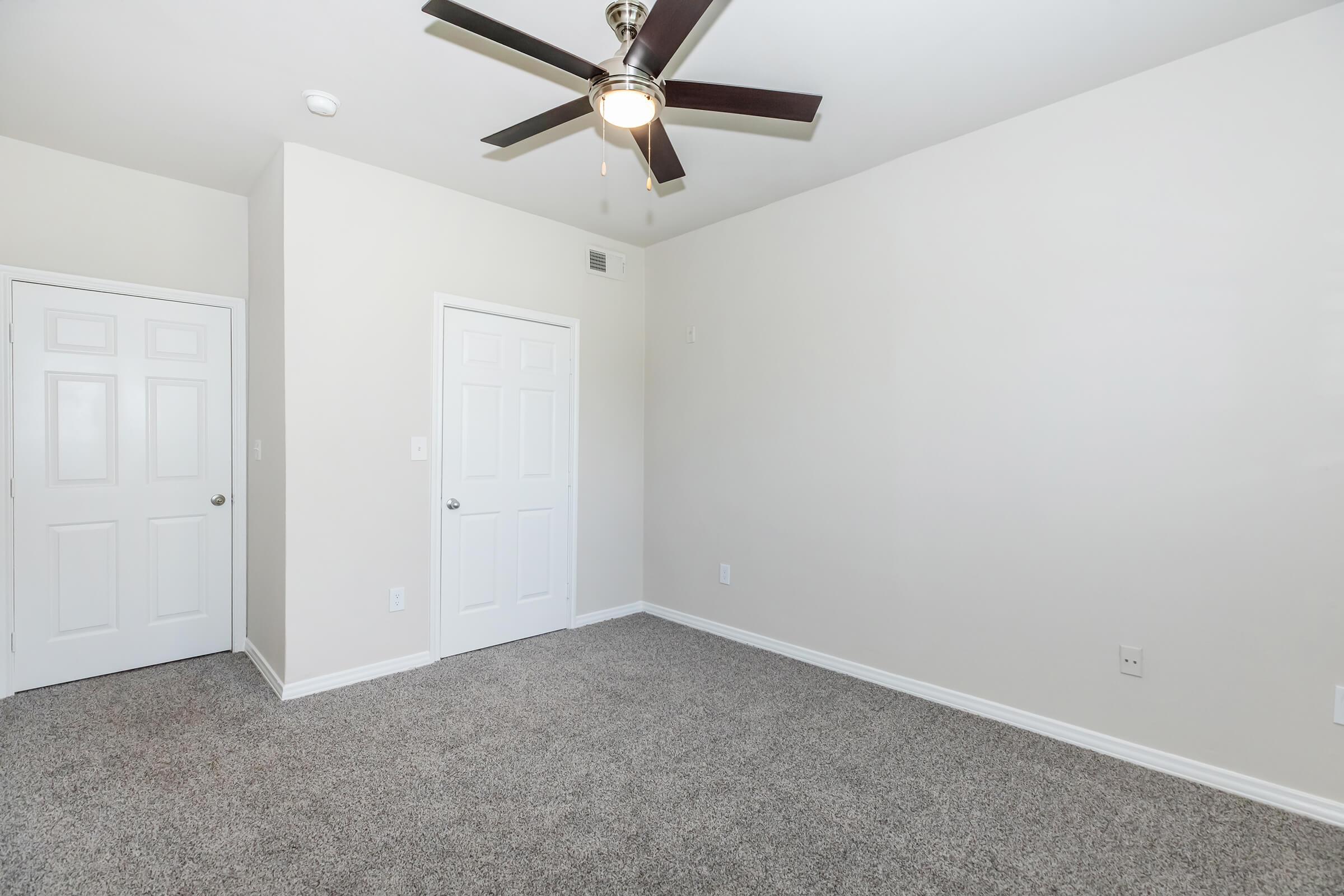 A clean, empty room featuring light beige walls, a ceiling fan, and gray carpet. There are two closed white doors on the left side, and the room is well-lit with natural light, creating a bright and spacious atmosphere.