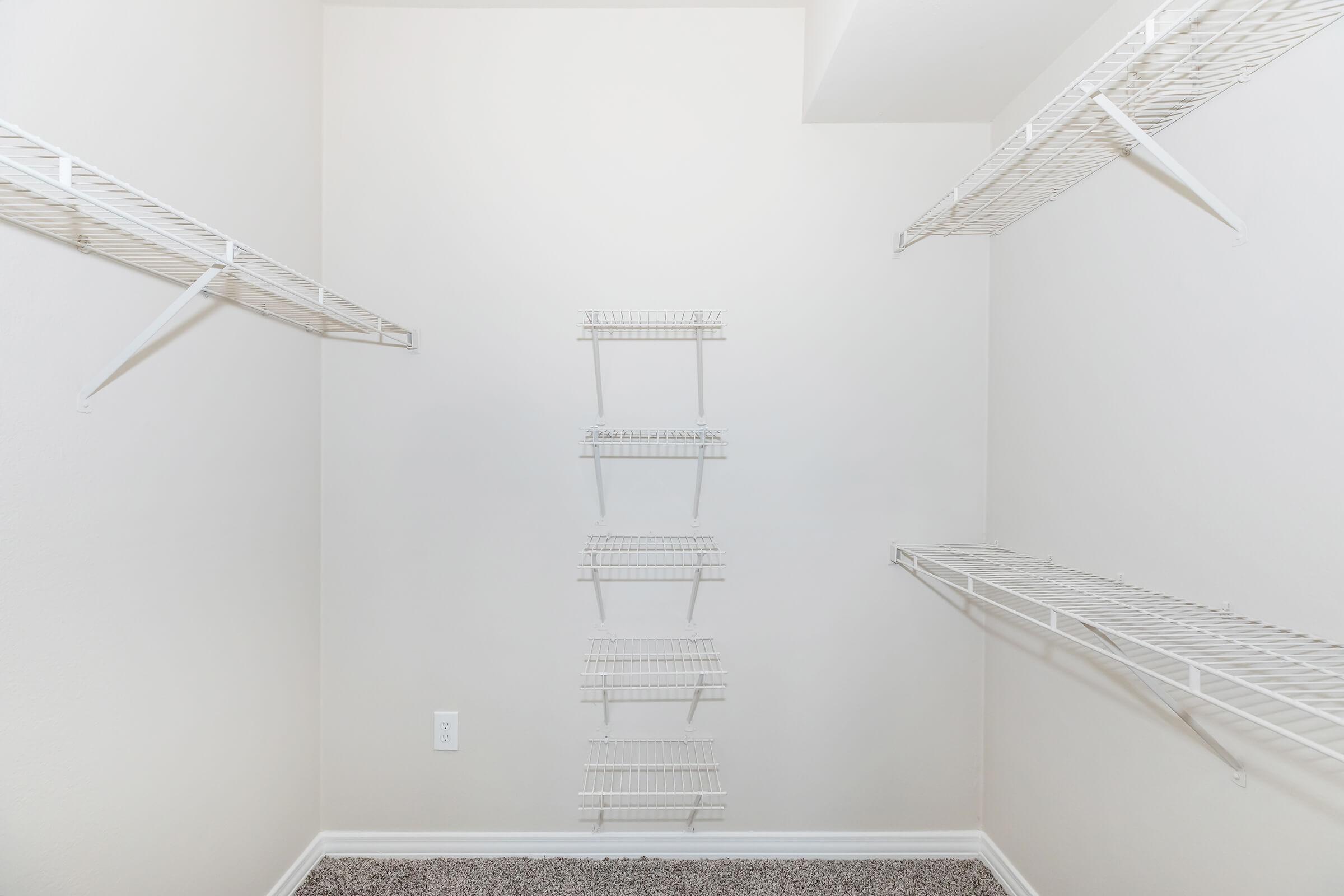 Empty closet with white wire shelves mounted on the walls, featuring five vertical shelves. The walls are painted a light color, and the floor is covered with gray carpeting. The overall space appears clean and organized, ready for storage.