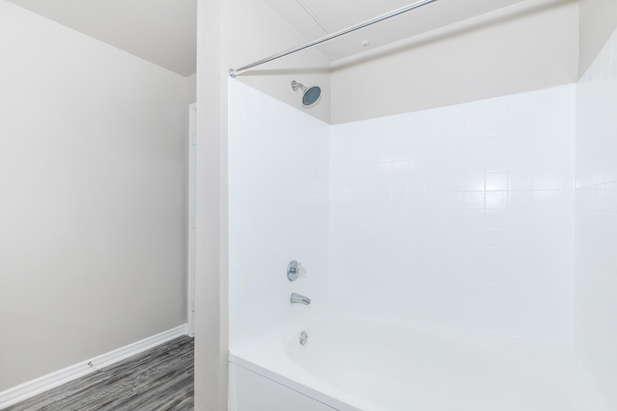A modern bathroom featuring a clean, white tiled shower with a curved glass partition. The shower includes a chrome showerhead and control knobs. The floor appears to be a light wood or laminate material, and the walls are painted in a neutral color.