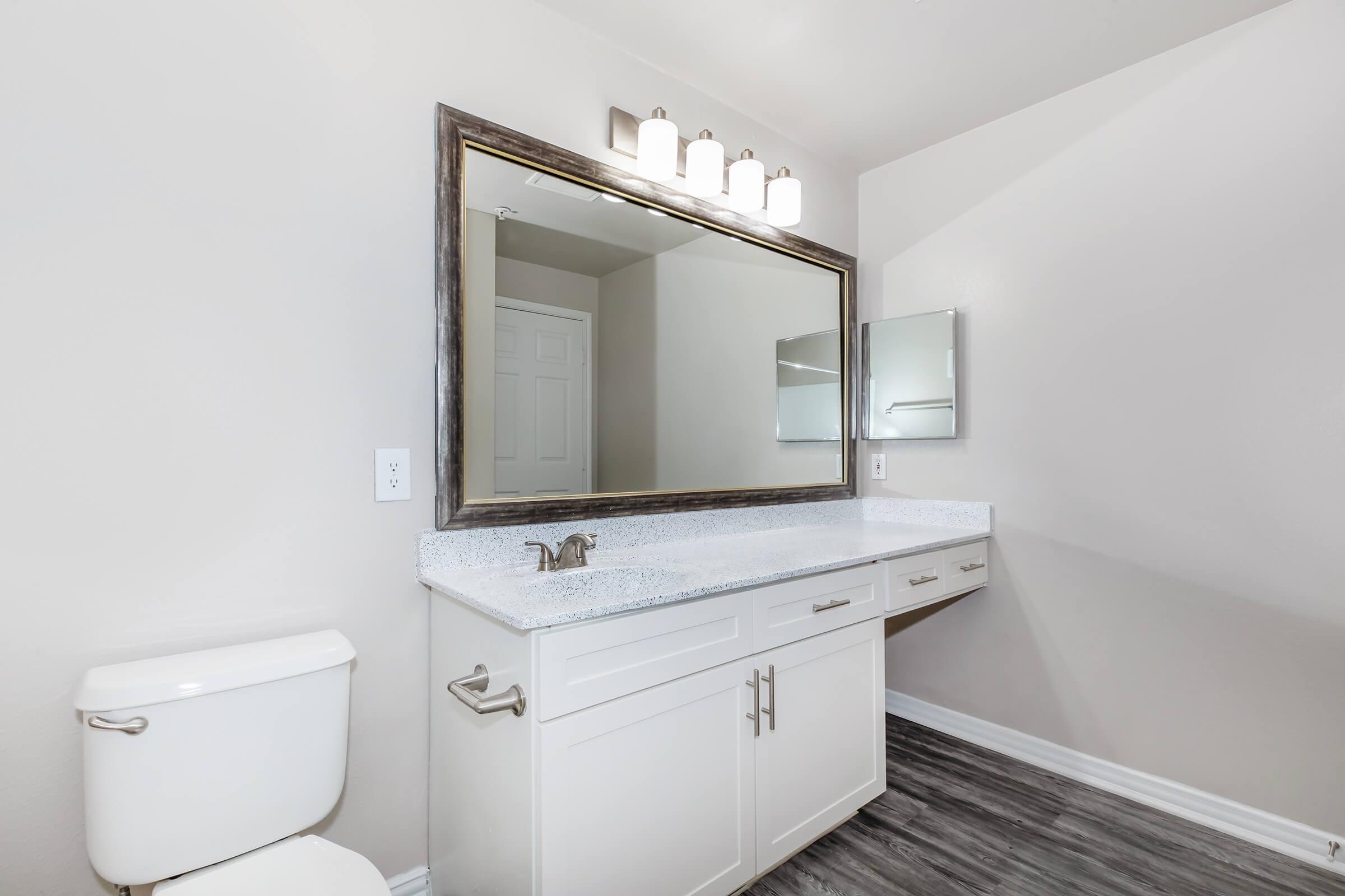 A modern bathroom featuring a white vanity with a granite countertop and mirror above. The walls are painted light gray, and there is a toilet to the left. The room has a sleek design with a large mirror and subtle lighting above. The floor showcases a wood-like finish.