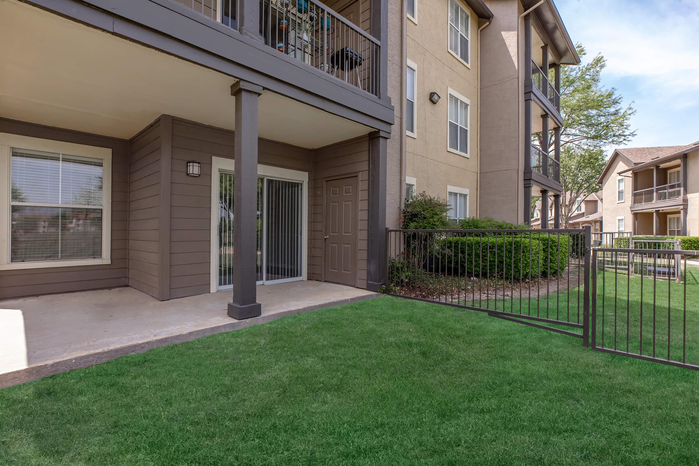 Ground-level view of a modern apartment exterior, featuring a patio area with a sliding glass door. The building has a neutral color scheme, surrounded by manicured green grass and fenced landscaping. Nearby, there are additional apartment units visible with balconies.