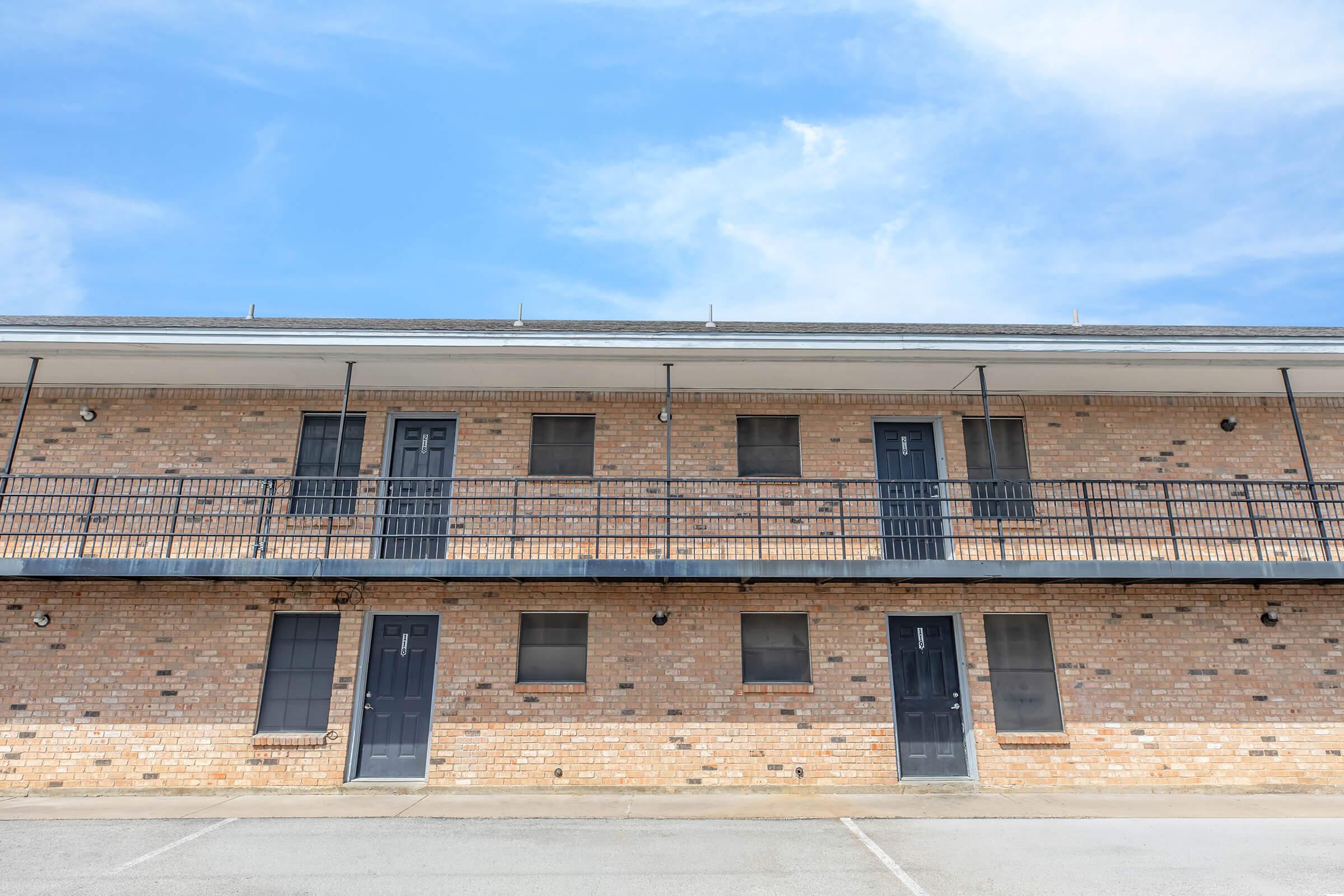 Two-story brick building with multiple entrances featuring double doors and balconies. The facade includes several windows, some with screens, on a clear day with a blue sky. The ground is paved, and there are no visible cars or people in the area.