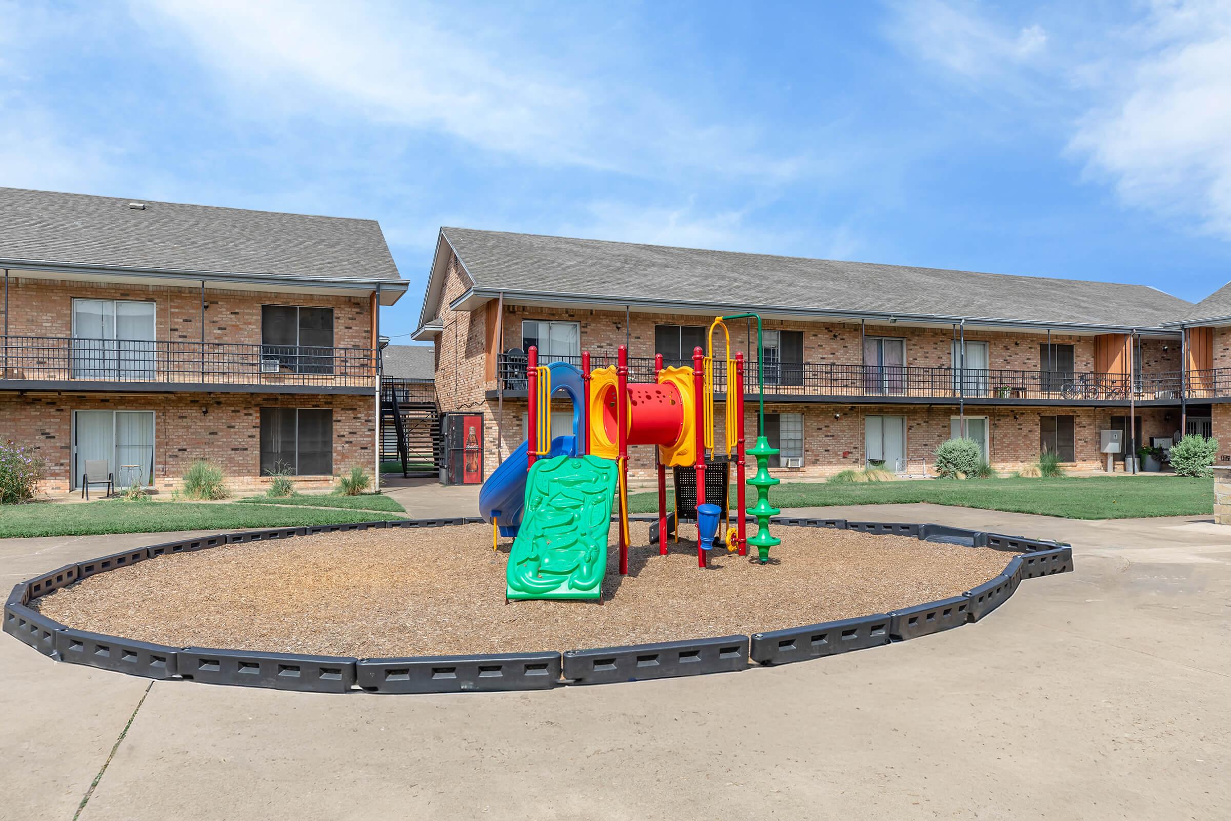 A colorful playground with a slide and climbing structure is situated in a grassy area surrounded by brick buildings. The scene features a clear blue sky, and the playground is designed for children, offering a safe space for outdoor play.
