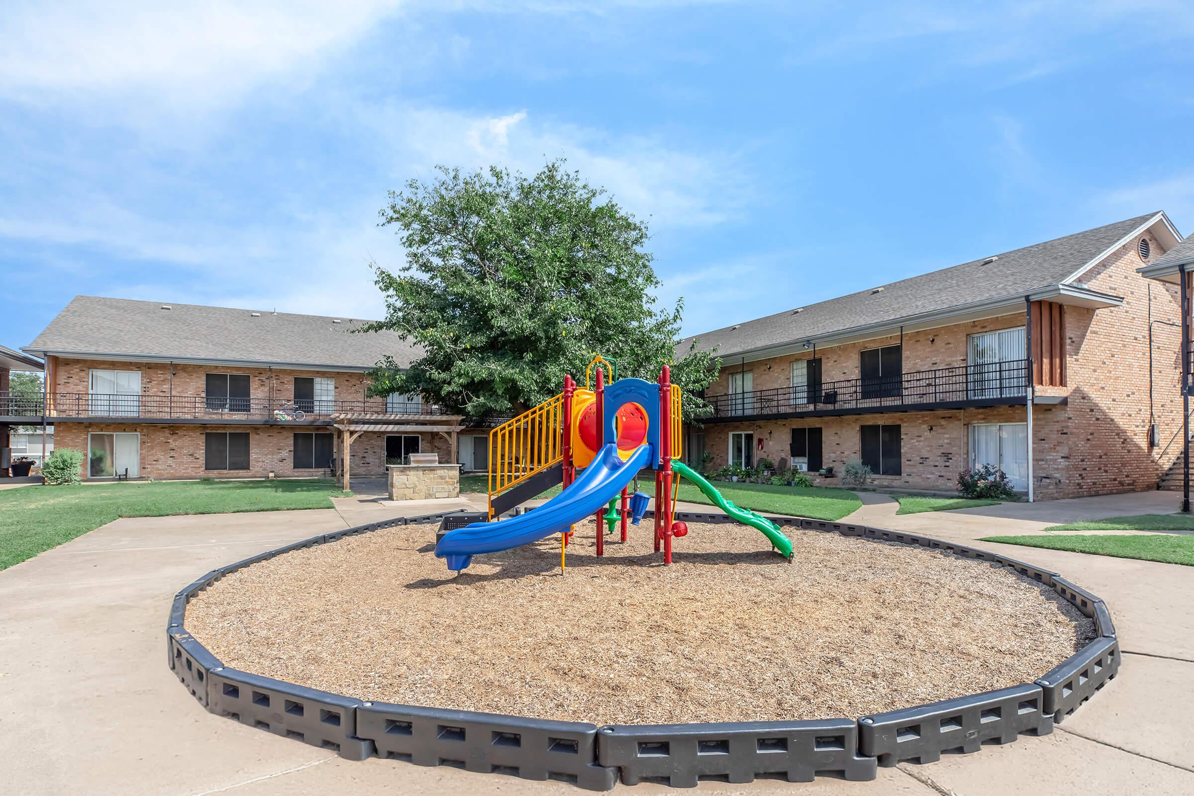 A playground area featuring a colorful slide and climbing structure, surrounded by gravel and enclosed by a black border. In the background, there are two brick apartment buildings with balconies, set against a clear blue sky. Green grass surrounds the playground, creating a vibrant outdoor space for children.