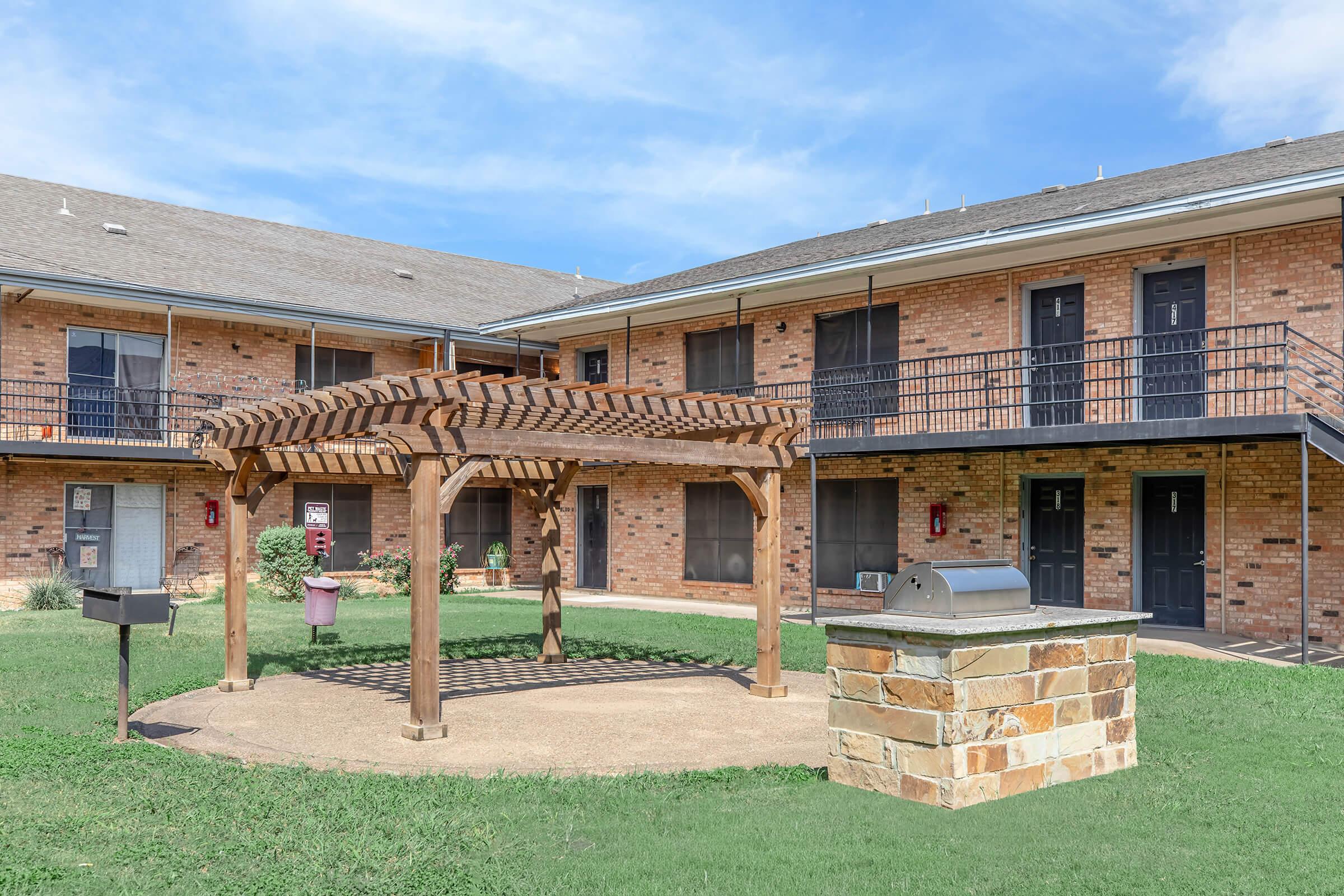 A courtyard area featuring a wooden pergola with seating, a stone grill, and a grassy lawn, surrounded by two brick apartment buildings with balconies and separate entrances. The sky is clear with a few clouds, creating a pleasant outdoor space.
