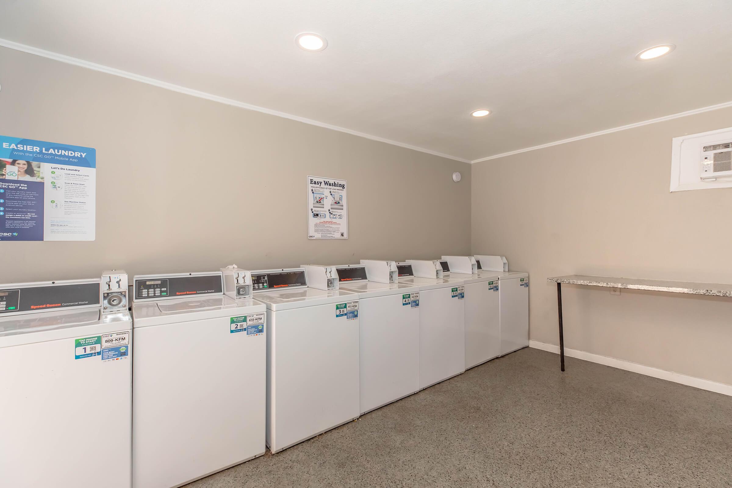 A bright laundry room featuring several white washing machines lined up against a beige wall. There's a folding table to the side and informational posters on the wall. The space is clean and well-lit, creating an inviting atmosphere for doing laundry.
