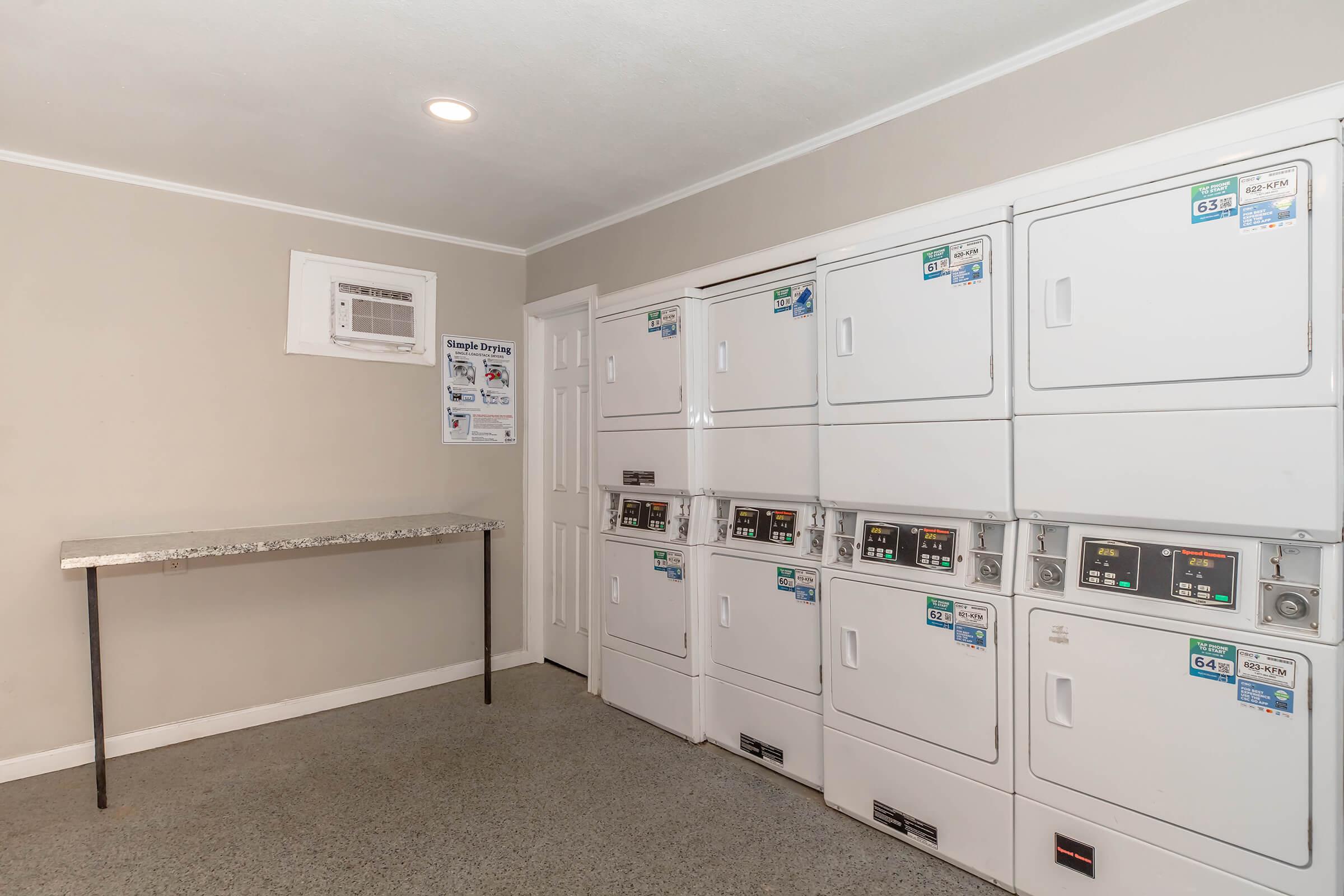 A clean, well-lit laundry room featuring multiple stacked washing machines and dryers, labeled with numbers. There's a small folding table in front of the machines. A window unit air conditioner is mounted on the wall, and a bulletin board is visible with information and notices.