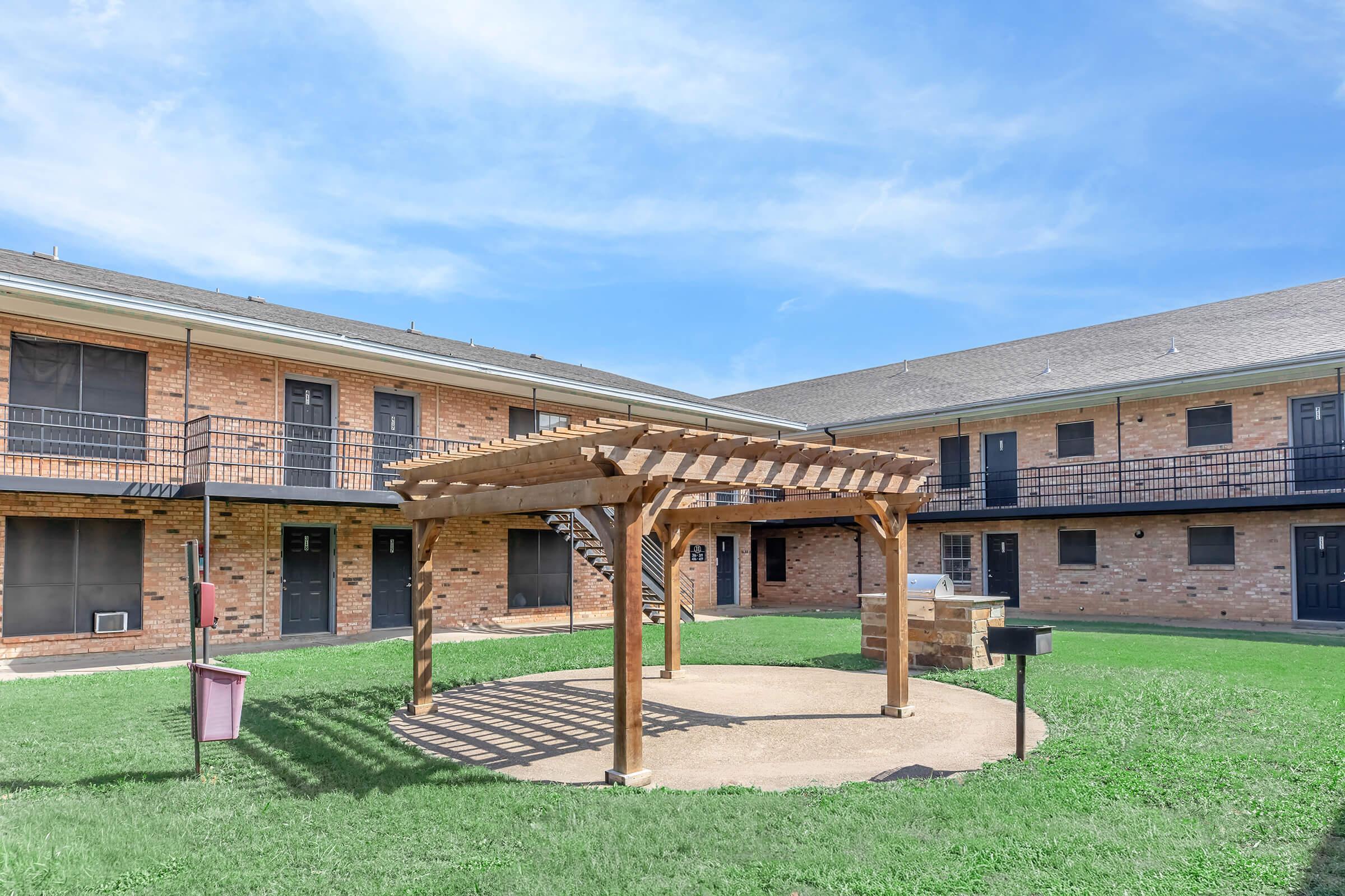 A courtyard featuring a wooden pergola with a circular seating area, surrounded by two-story brick buildings with multiple doors and balconies. Green grass is visible, along with a grill station and a clear blue sky above.