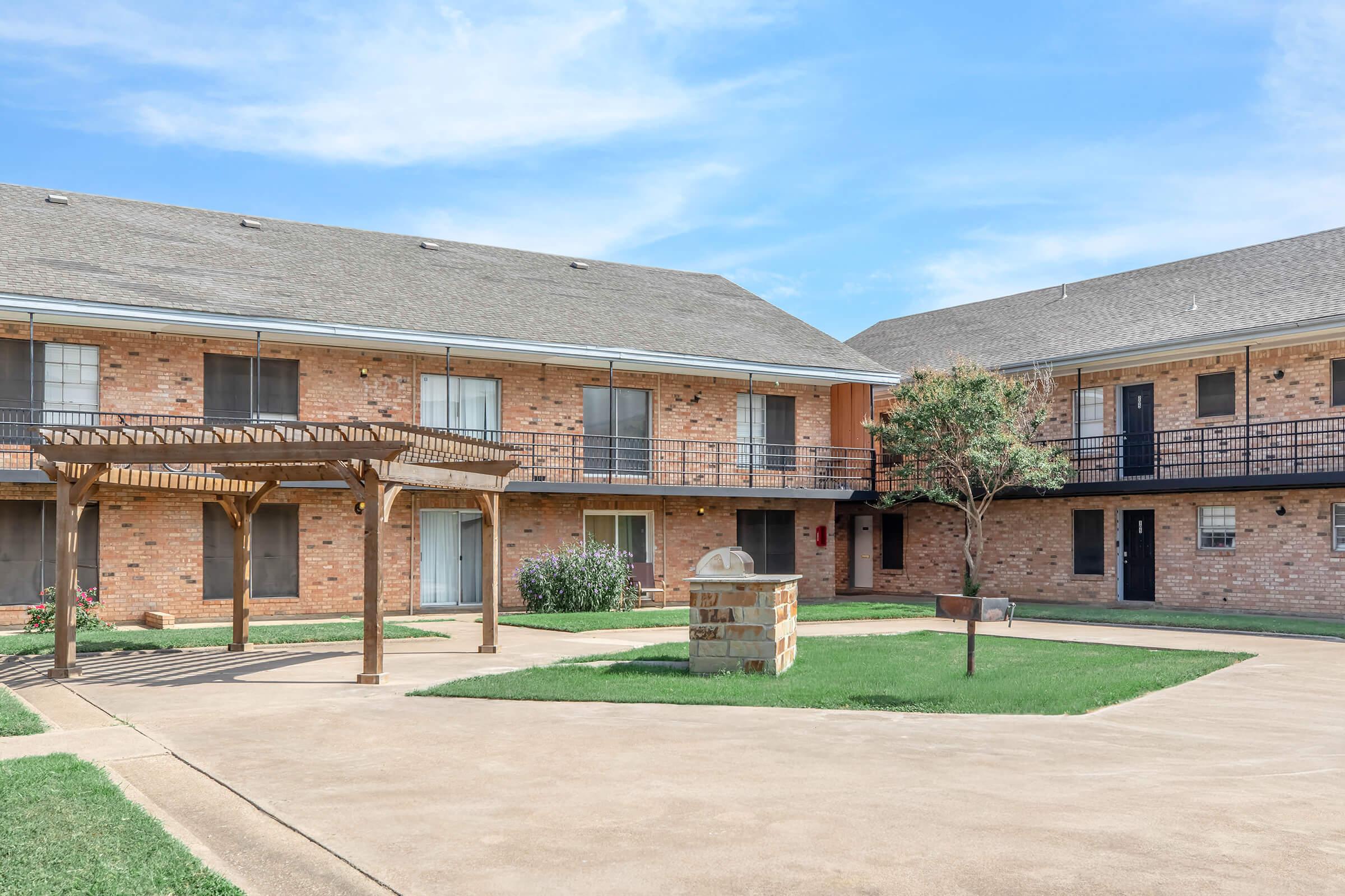 A two-story brick apartment building featuring a central courtyard with grass and a decorative stone sign. A wooden pergola is situated in the foreground, and there are multiple balconies and windows visible on the building's exterior. The sky is clear with a few clouds.
