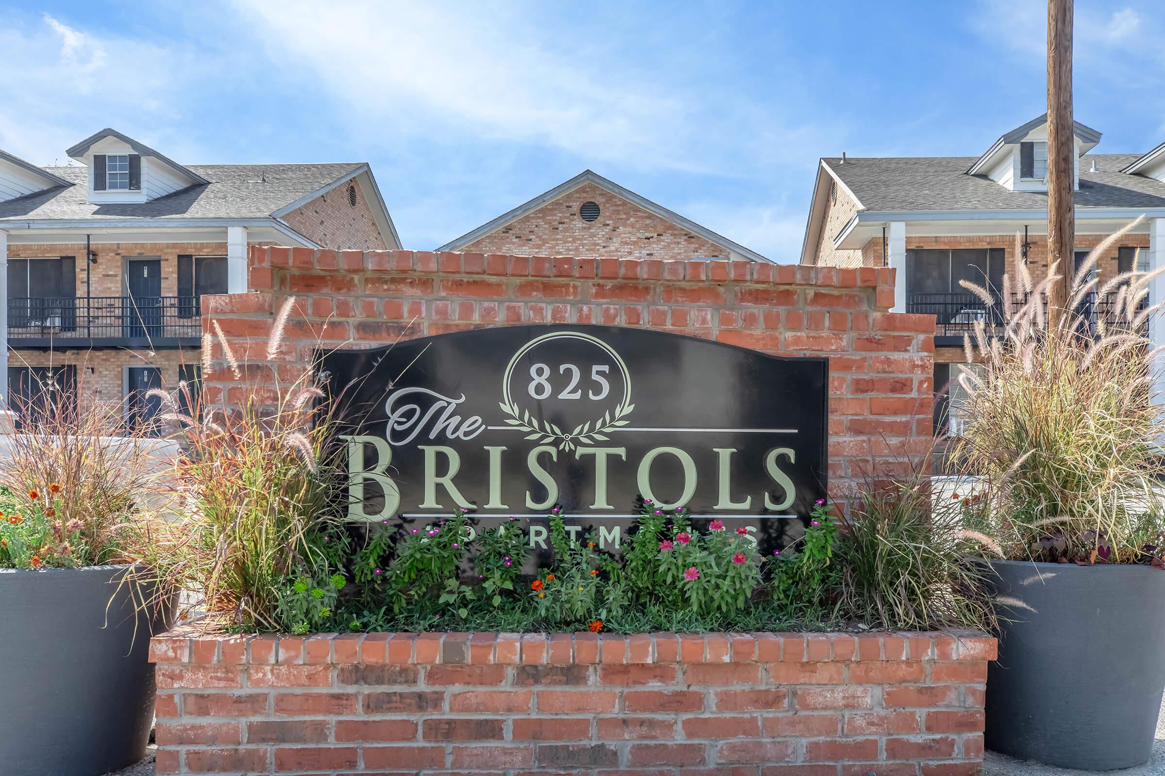 Sign for "The Bristols Apartments" at 825, with a brick base and decorative landscaping featuring flowers. The background shows a multi-story brick apartment building under a clear blue sky.