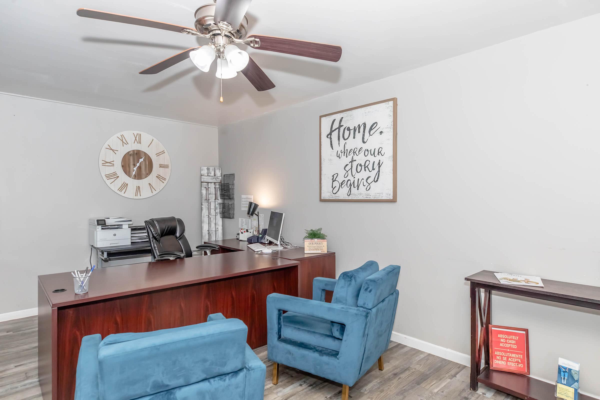 A cozy office space featuring a wooden desk with a computer, a printer, and decorative items. Two blue upholstered chairs are positioned in front. A wall clock and a framed motivational quote that reads "Home. Where our story begins" add character to the neutral-toned room. A small bookshelf displays a few books.