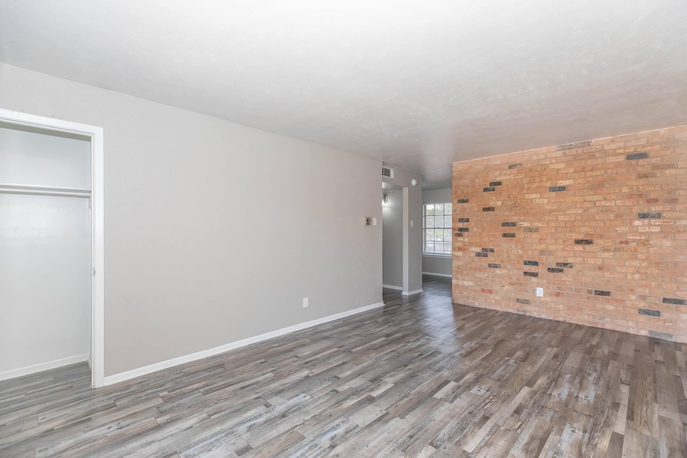 A spacious living area featuring light gray walls, a brick accent wall with black and tan bricks, and large, light-colored wooden flooring. A closet is visible on the left, and a window lets in natural light from an adjacent room.