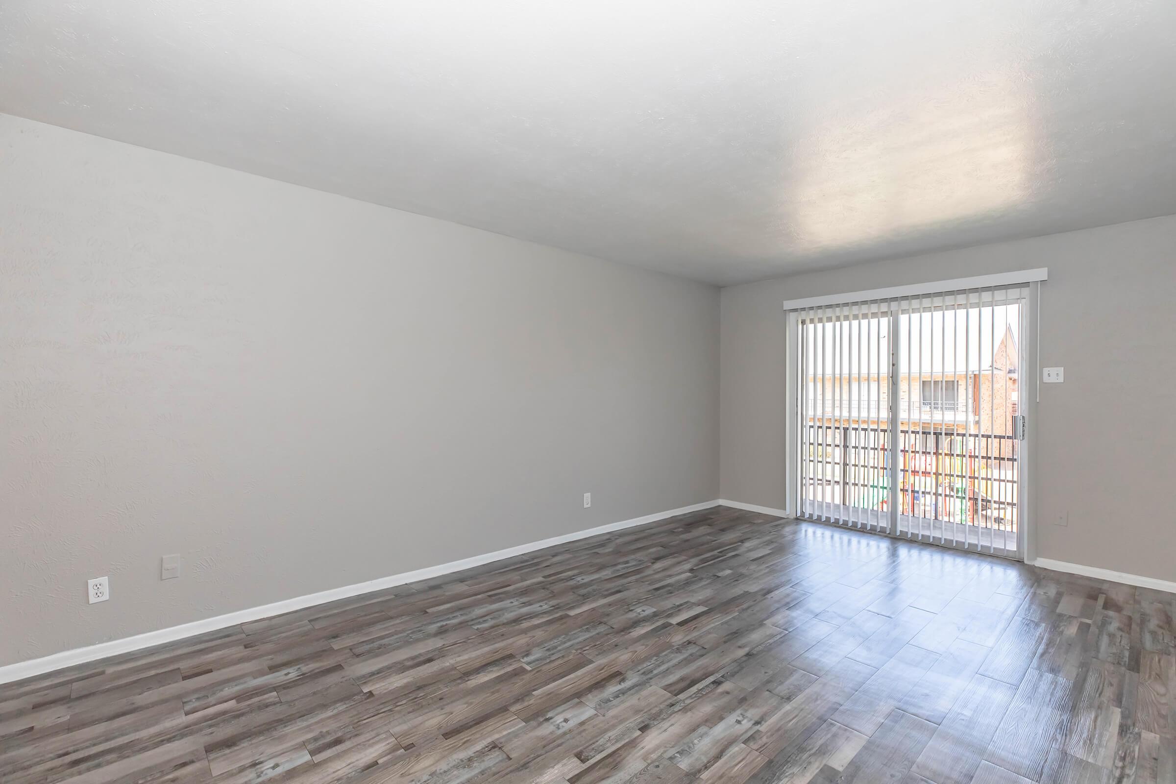 A spacious, empty living room with light gray walls and large windows. The floor features wooden laminate planks, and there is a sliding glass door leading to a balcony. Natural light fills the room, creating a bright and airy atmosphere.