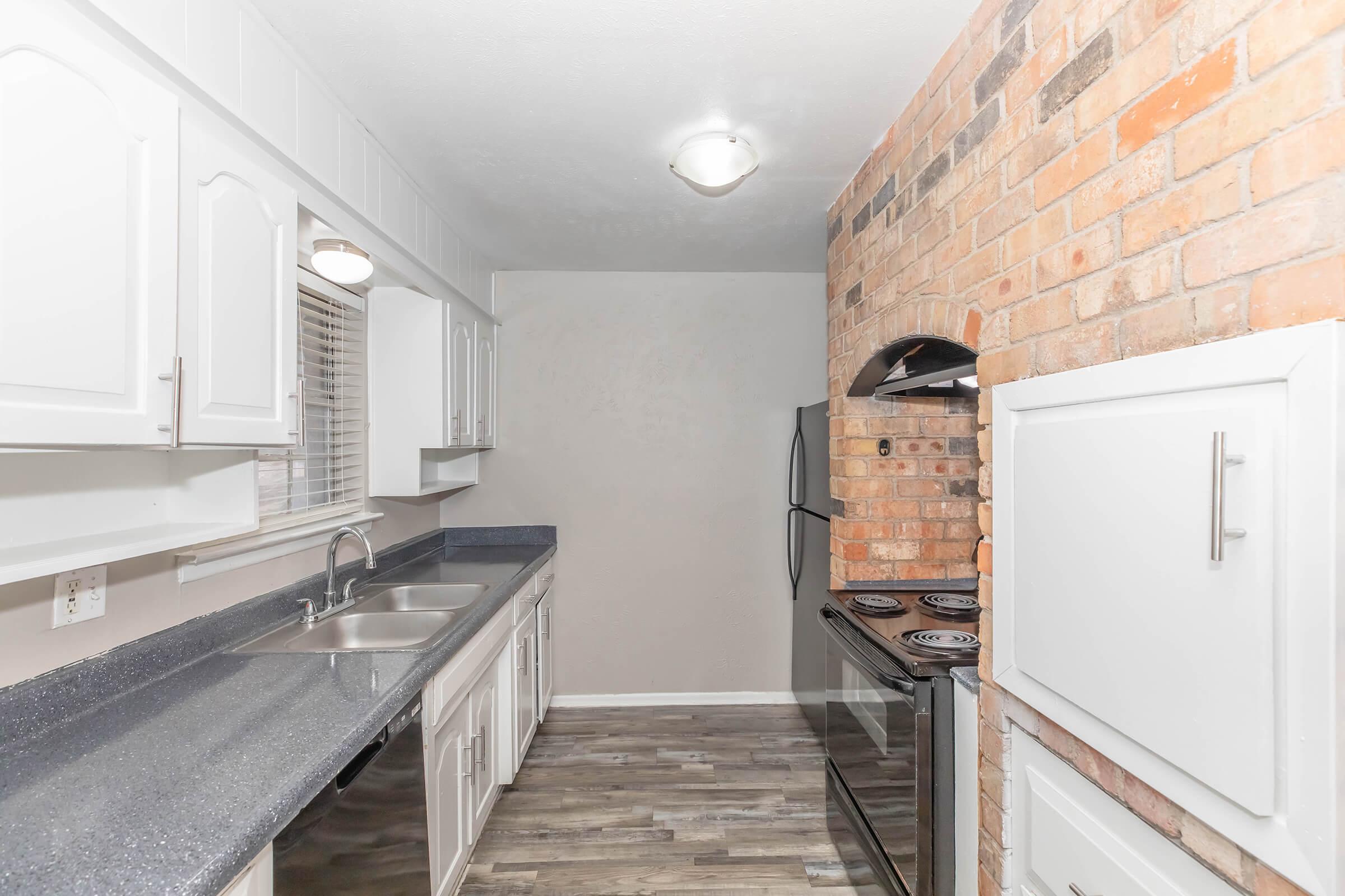 A modern kitchen featuring white cabinets, a gray countertop, stainless steel appliances, and a brick accent wall. The layout includes a double sink, an electric stove, and ample storage space. The flooring consists of laminated planks, providing a contemporary look.